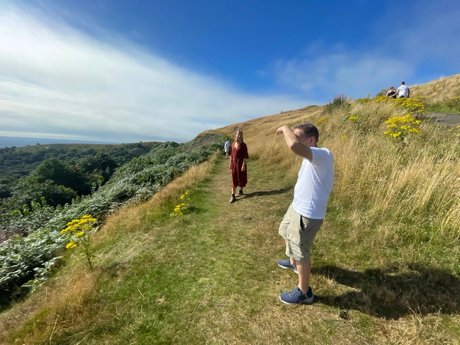 People walking along a hilltop trail in the Malverns with greenery, yellow flowers, and a view of rolling hills under a blue sky with clouds.