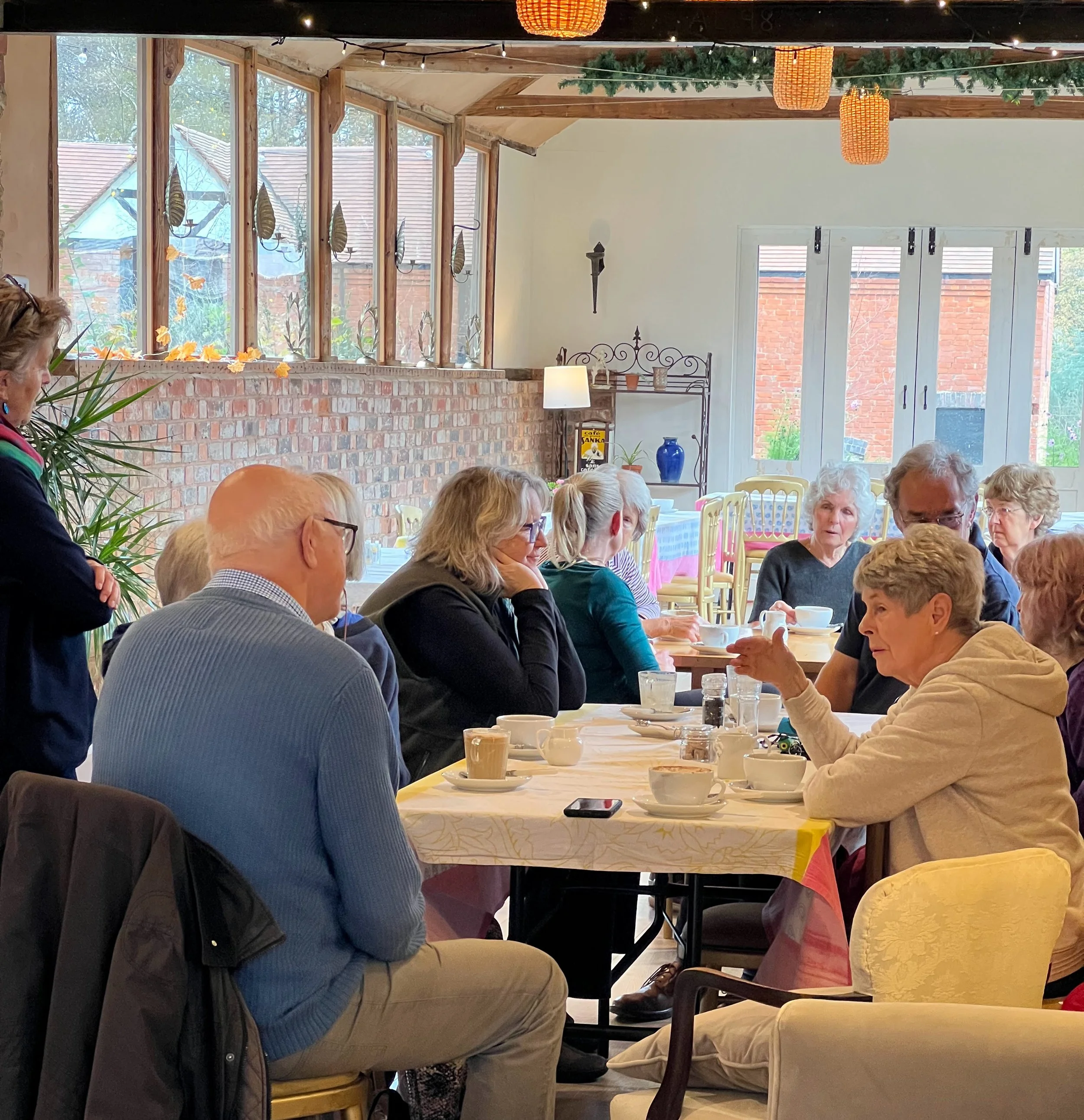 Group of elderly people sitting around a table in a cozy, decorated restaurant or cafe, engaged in conversation. Sunlight streams through large windows, with brick walls, hanging lights, and seasonal decorations visible in the background.