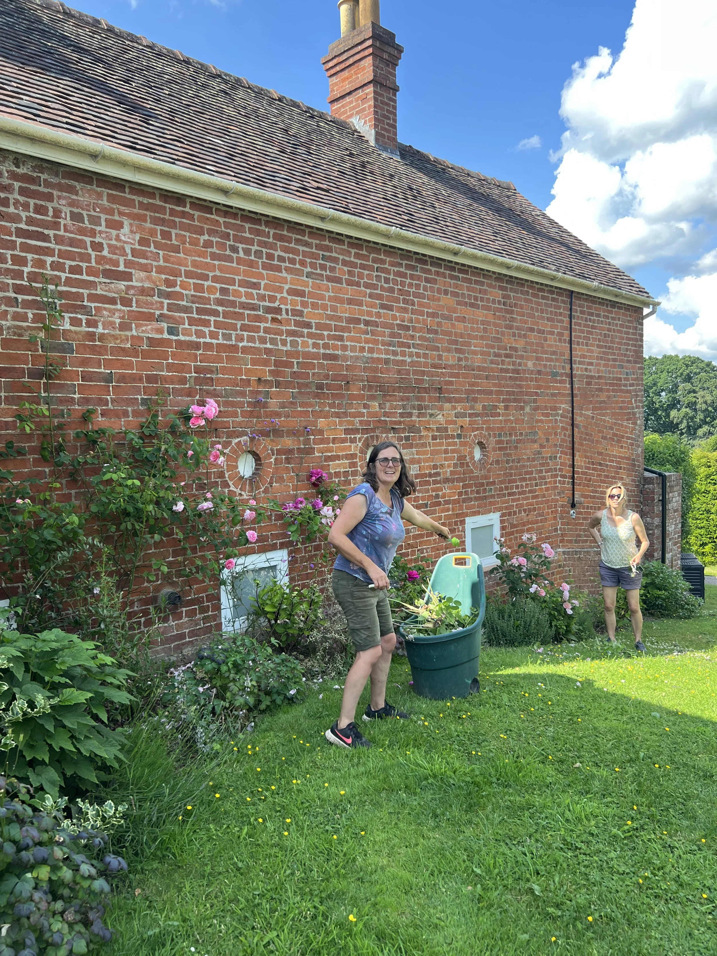 Two women outdoors in a garden next to a brick house with flowering bushes, one woman is handling garden waste with a trash bin, another woman is standing nearby. Clear sky with some clouds.