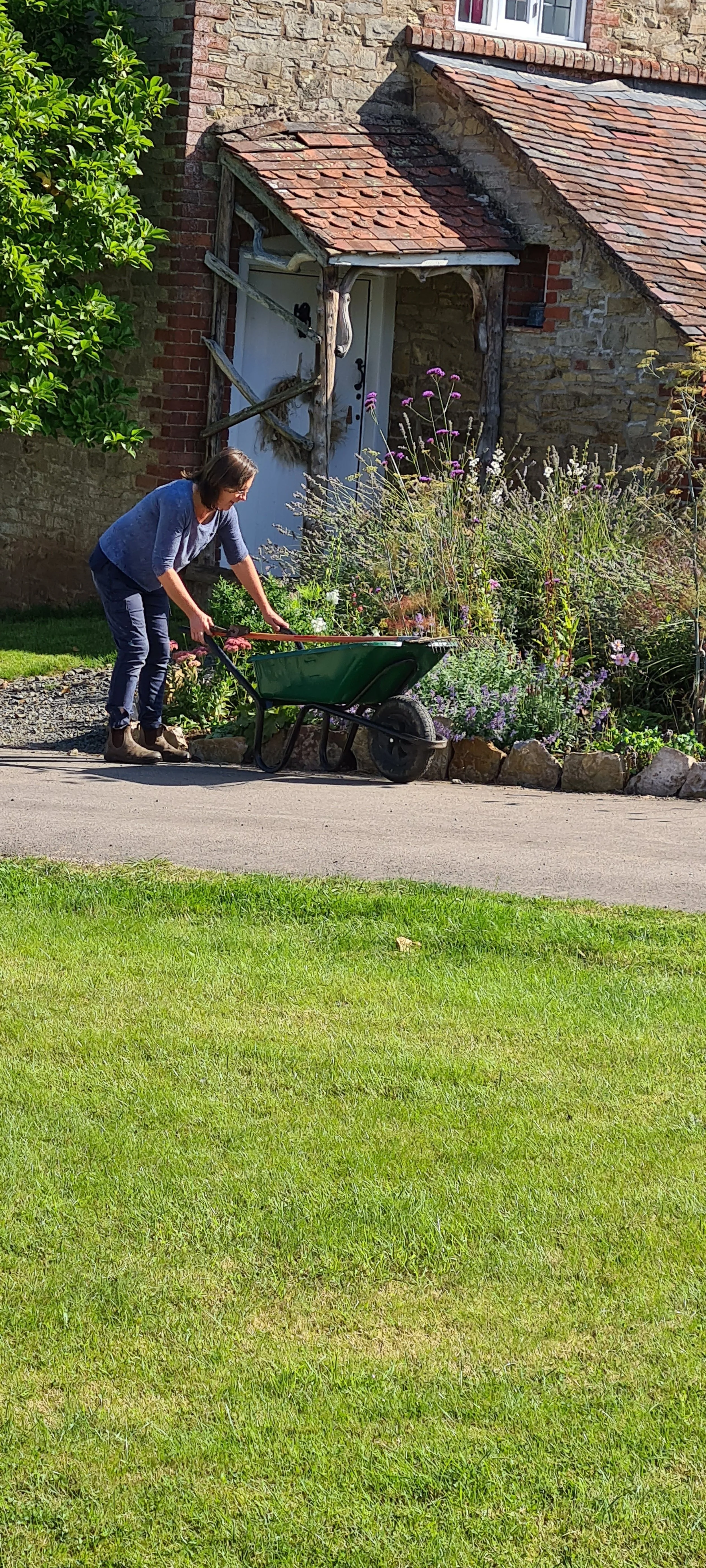 Woman pushing a green wheelbarrow along a garden path next to a colorful garden with flowering plants and a rustic brick and stone house in the background.