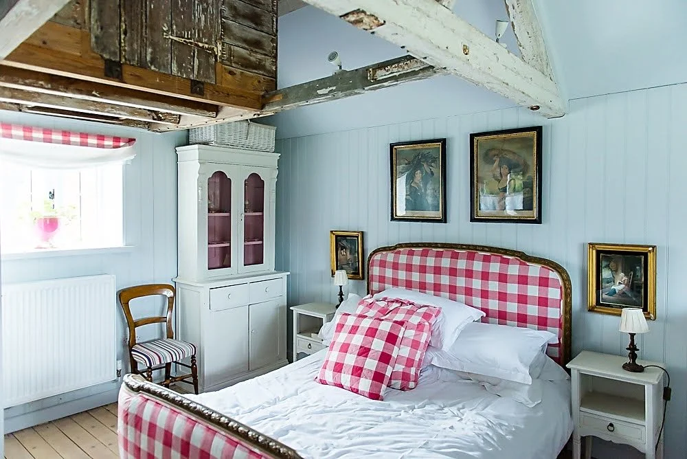 A cozy bedroom with a bed featuring a pink and white checkered headboard and matching pillow. There are white nightstands with lamps on each side of the bed. A white cabinet with glass doors is against the wall, and a wooden chair with a striped cush