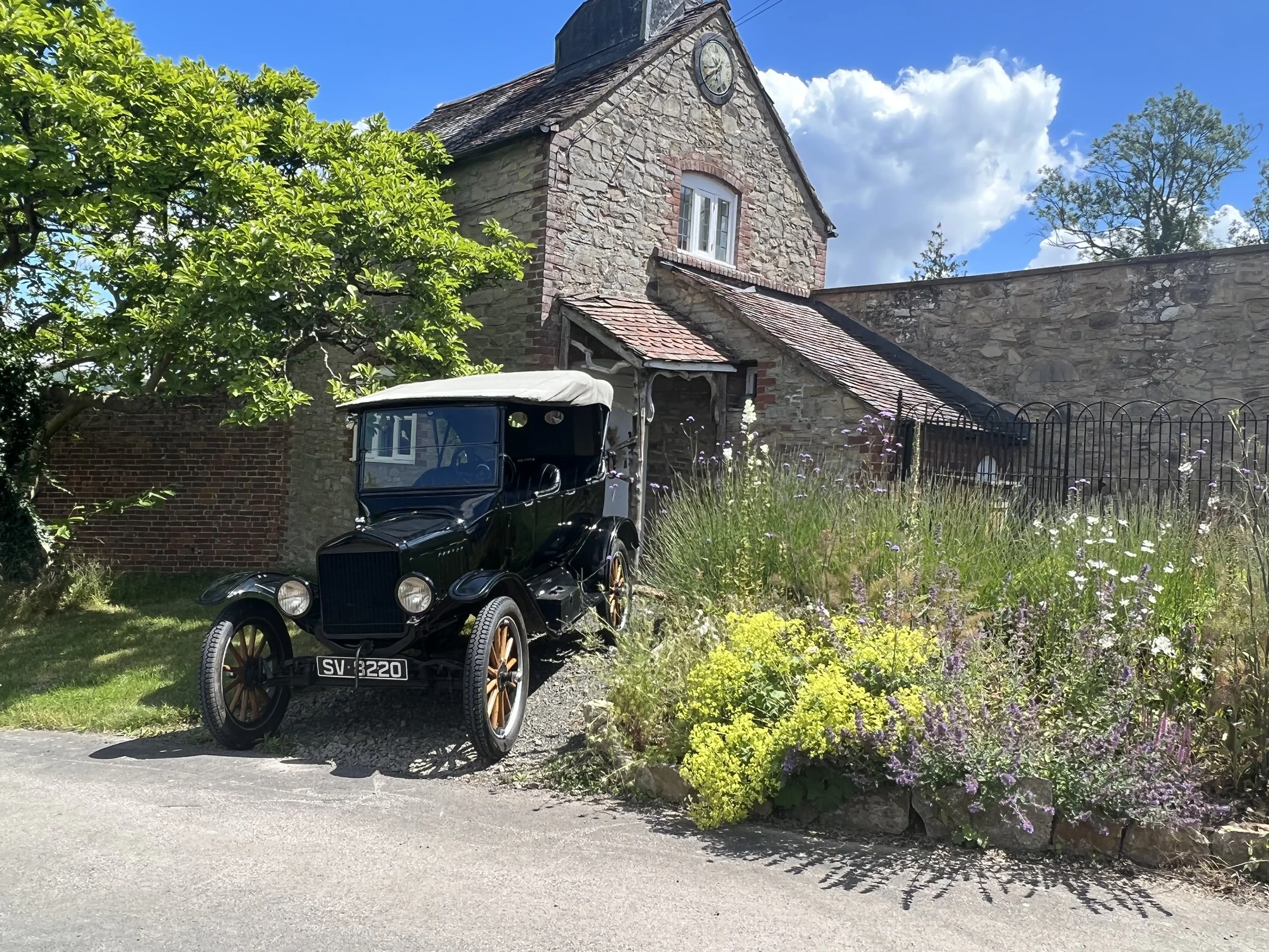 A vintage black car parked in front of a stone building with a clock on its gable in a garden with a variety of flowers and a green tree.