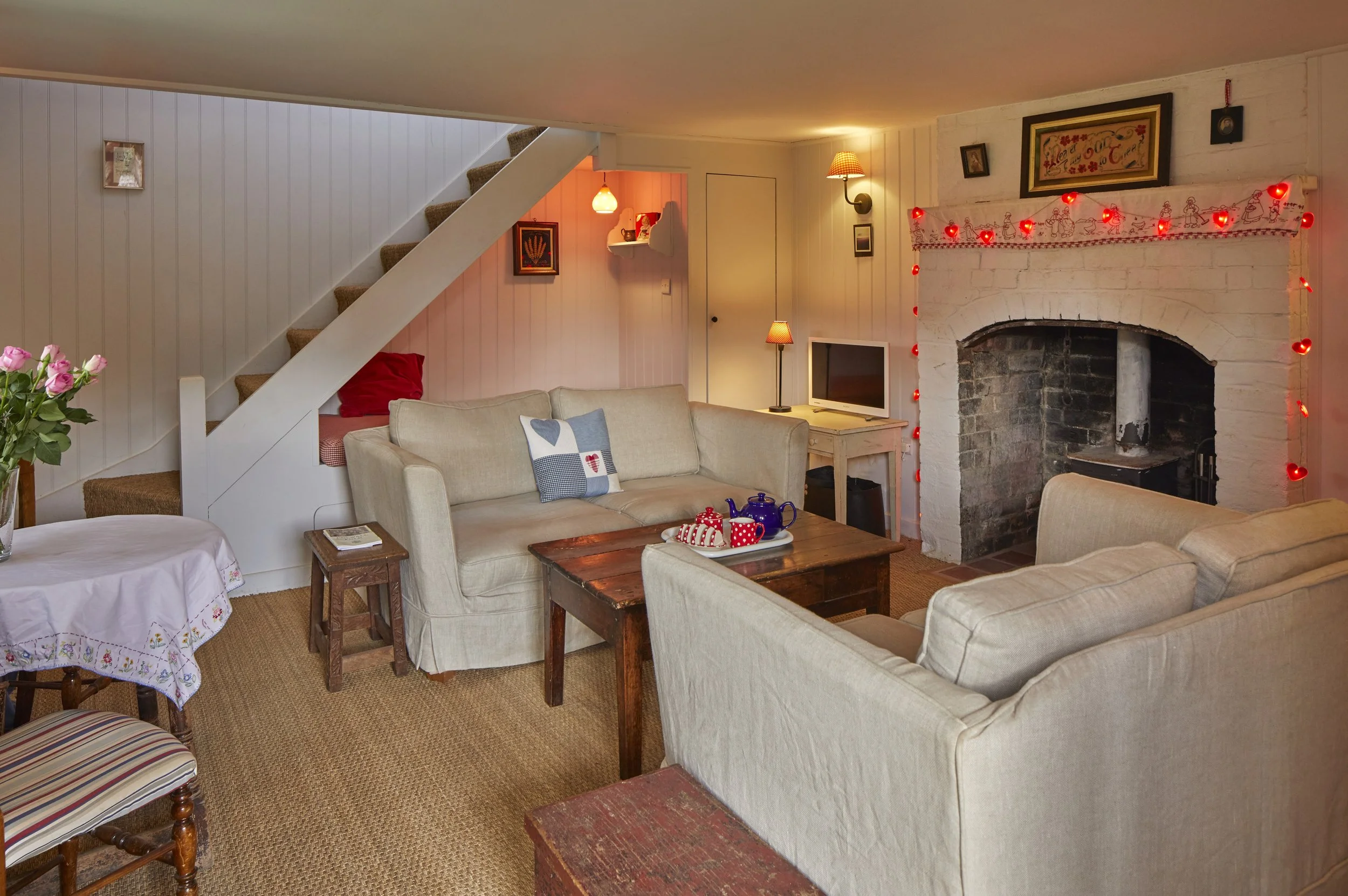 Cozy living room with beige couches, a fireplace decorated with red string lights, a small TV, a wooden coffee table with blue and red teapots and cups, a round table with pink roses, and wall art, with a staircase leading upstairs.