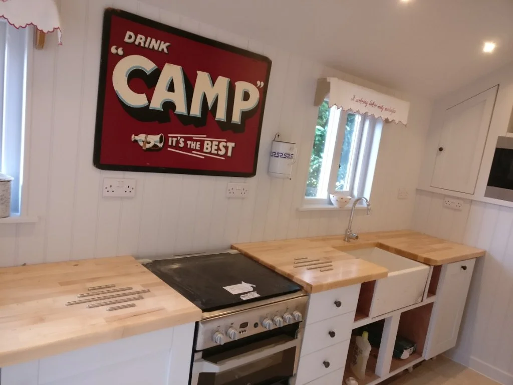 Kitchen with wooden countertops, white cabinets, a stove, a small window above the sink, and a large red sign that reads "Drink CAMP, It's the Best."
