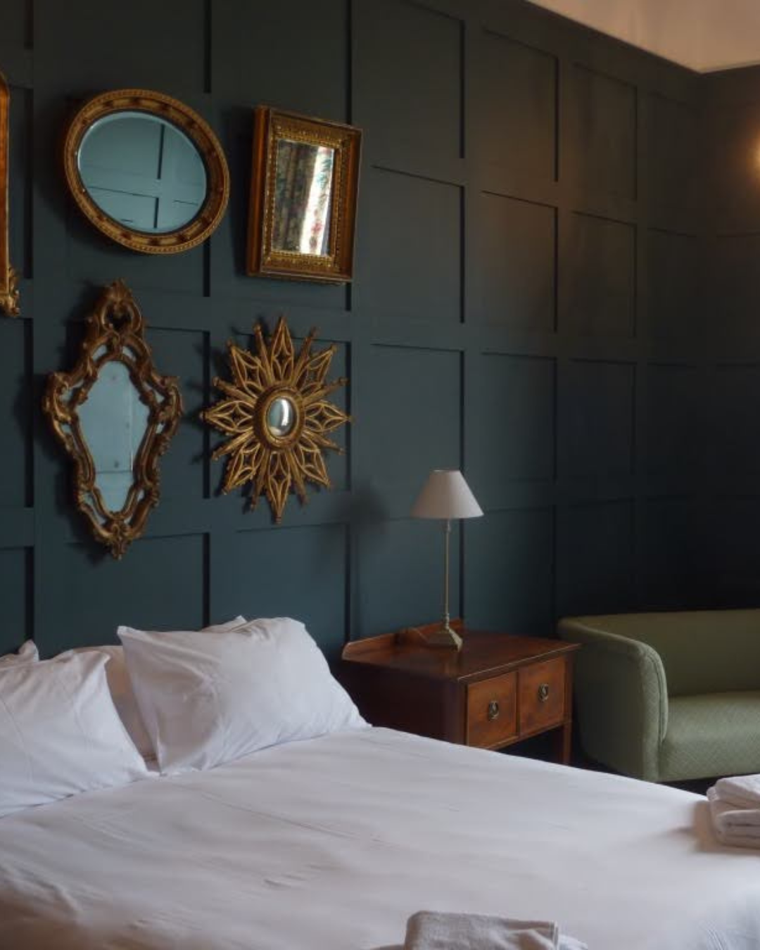 A cozy bedroom with a bed, wooden nightstand, and a green armchair. The dark blue wall behind the bed is decorated with several ornate, vintage-style mirrors and picture frames, with a lamp on the nightstand.