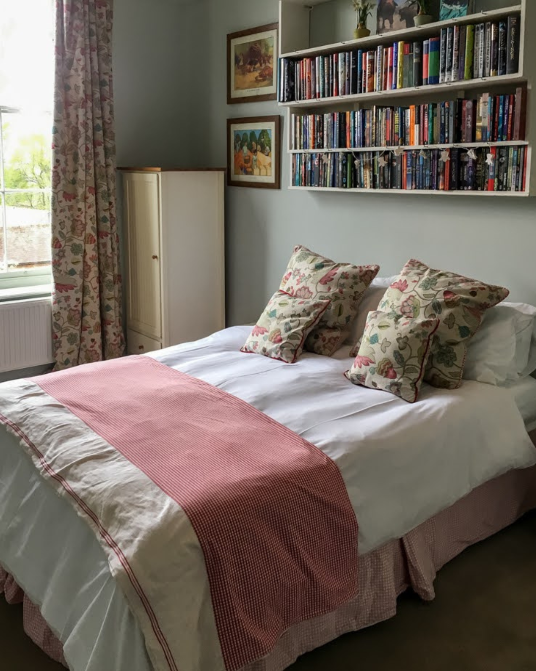 A neatly made bed with white sheets, three floral pillows, and a pink patterned bed runner in a bedroom with a window, floral curtains, a white cabinet, and a white bookshelf filled with books on the wall.