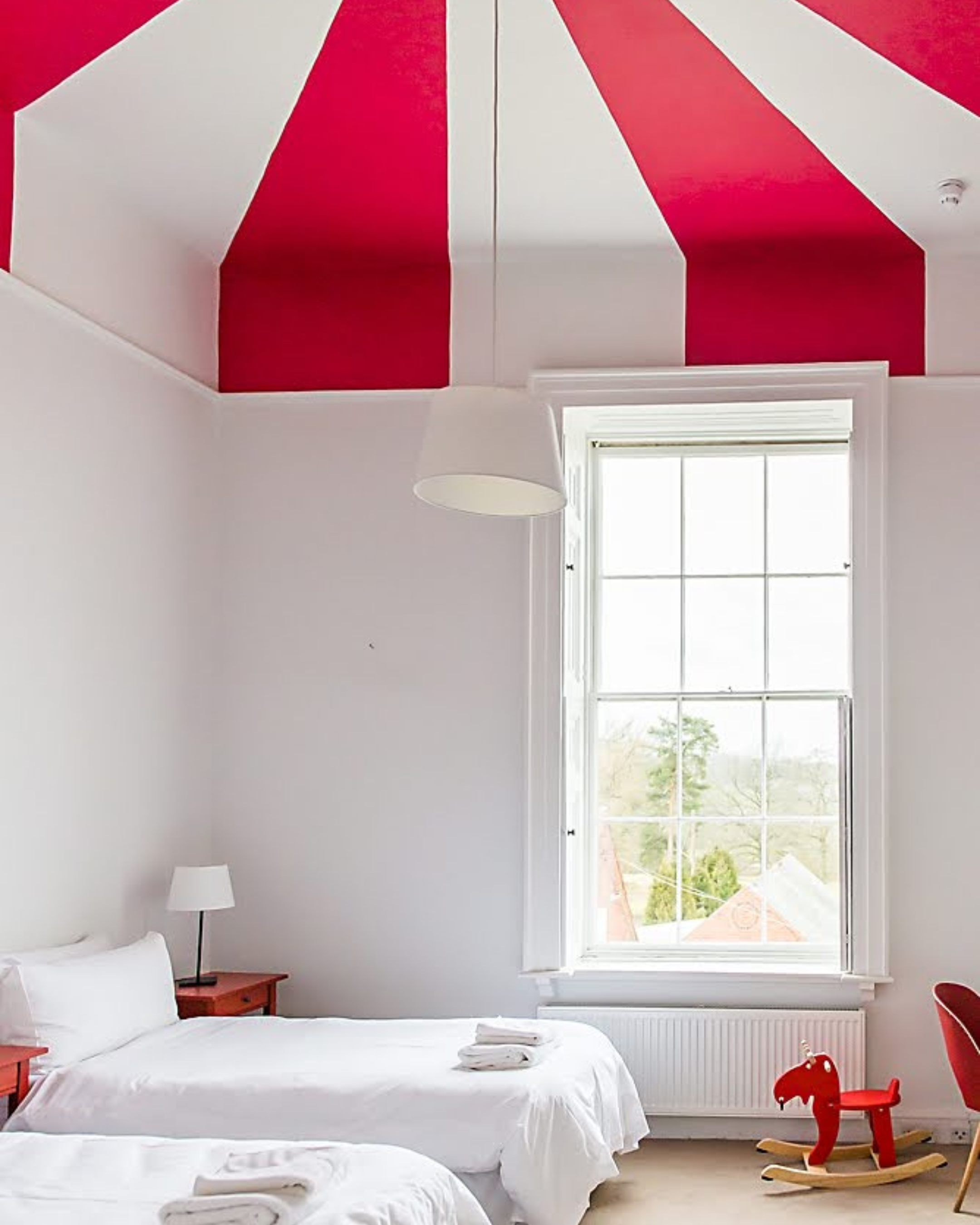 A bright bedroom with two twin beds, white bedding, and towels on each bed. There are two red nightstands with lamps, a large window with white trim, a red and white striped ceiling, and a red rocking horse near a red chair.