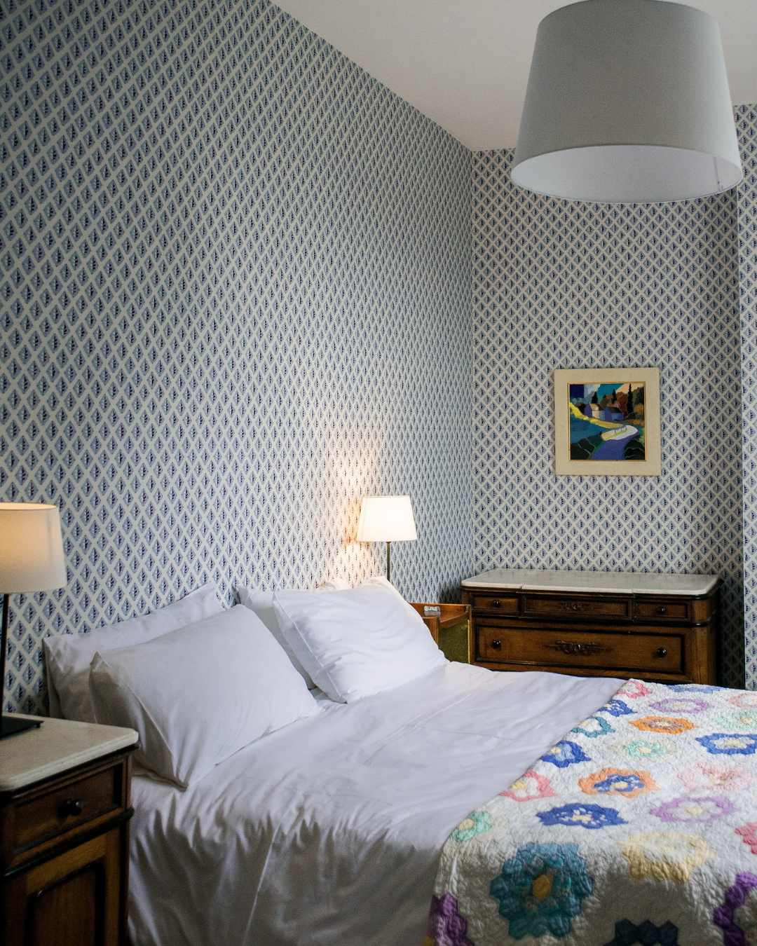 A bedroom with a bed, white pillows, and a colorful quilt. The walls have blue and white diamond-patterned wallpaper. There are two lamps, one on each side of the bed, and a framed landscape painting on the wall above a wooden dresser.