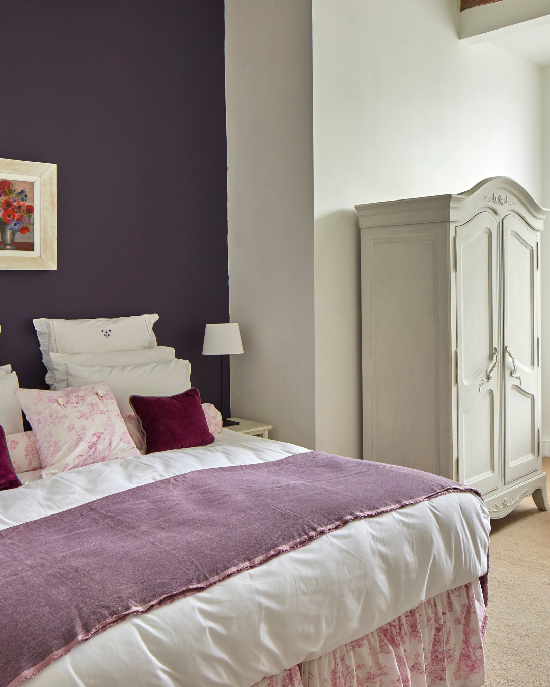 A bedroom with a purple accent wall and a white bed with pink and purple pillows and a pink bedspread. There's a white wardrobe and a small nightstand with a lamp.