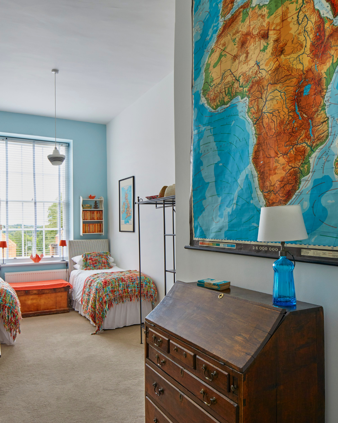 A bedroom with two beds, colorful blankets, a large window, a map of Africa on the wall, and vintage furniture.