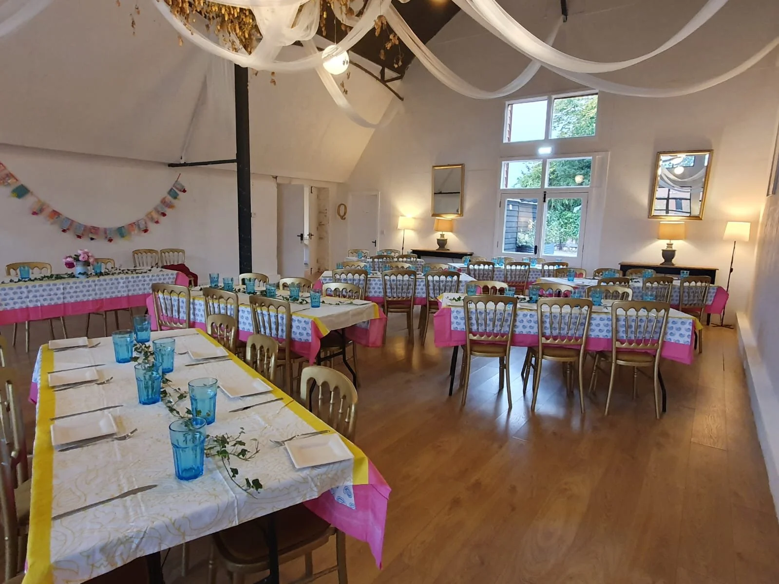 An indoor celebration space decorated with vintage-style tables and chairs, with pink and yellow accents, and a chandelier with white fabric drapes hanging from the ceiling.