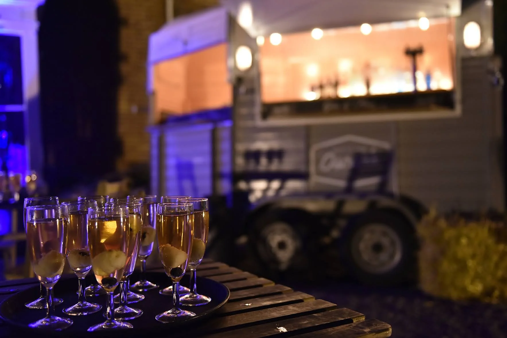 Several glasses of champagne with olives on a wooden table at an outdoor party at night, with a mobile bar trailer in the background illuminated with warm lights.