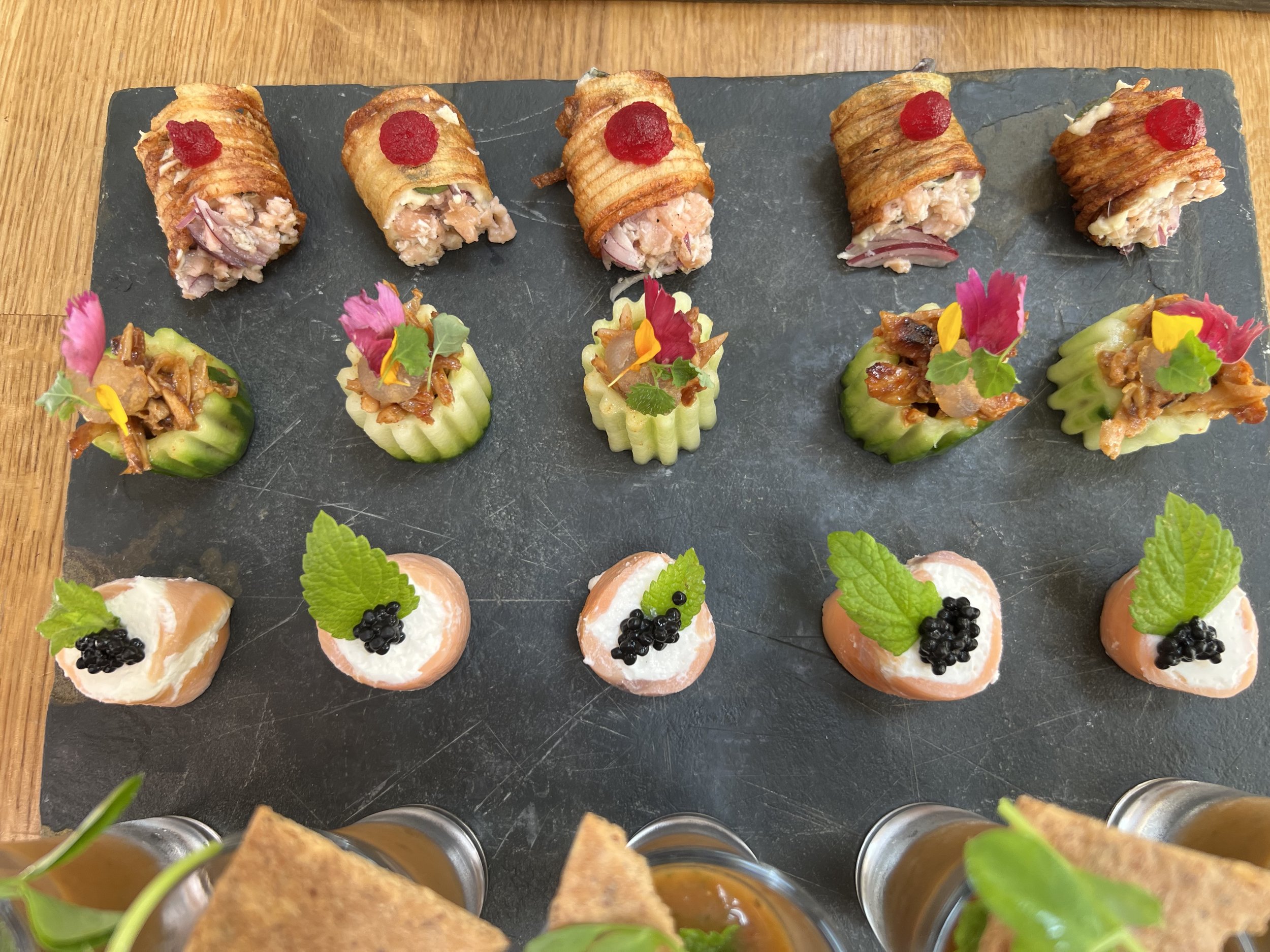 A platter of assorted appetizers with three rows of bite-sized foods on a dark slate serving tray, including rolled and stuffed pastries, cucumber cups with toppings, and small heart-shaped salmon bites garnished with herbs and caviar.
