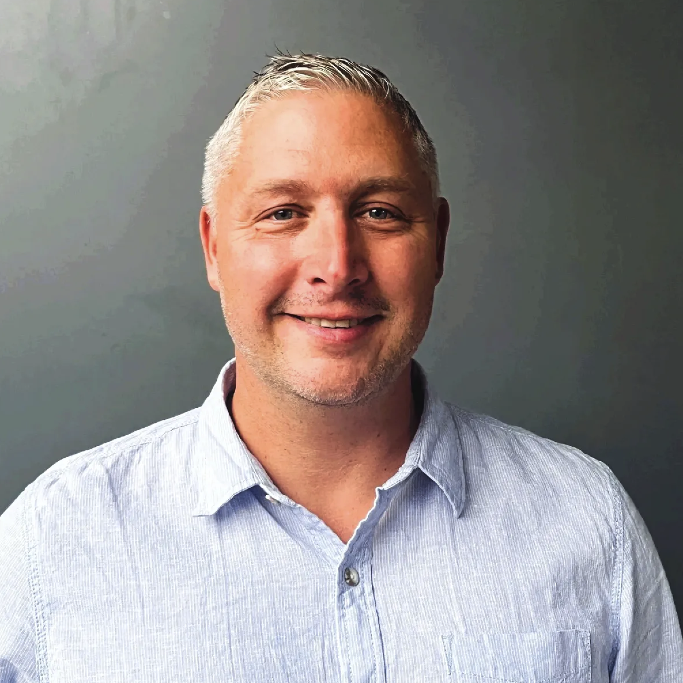 Portrait of a smiling man with short blonde hair, wearing a light blue collared shirt, standing against a plain gray background.