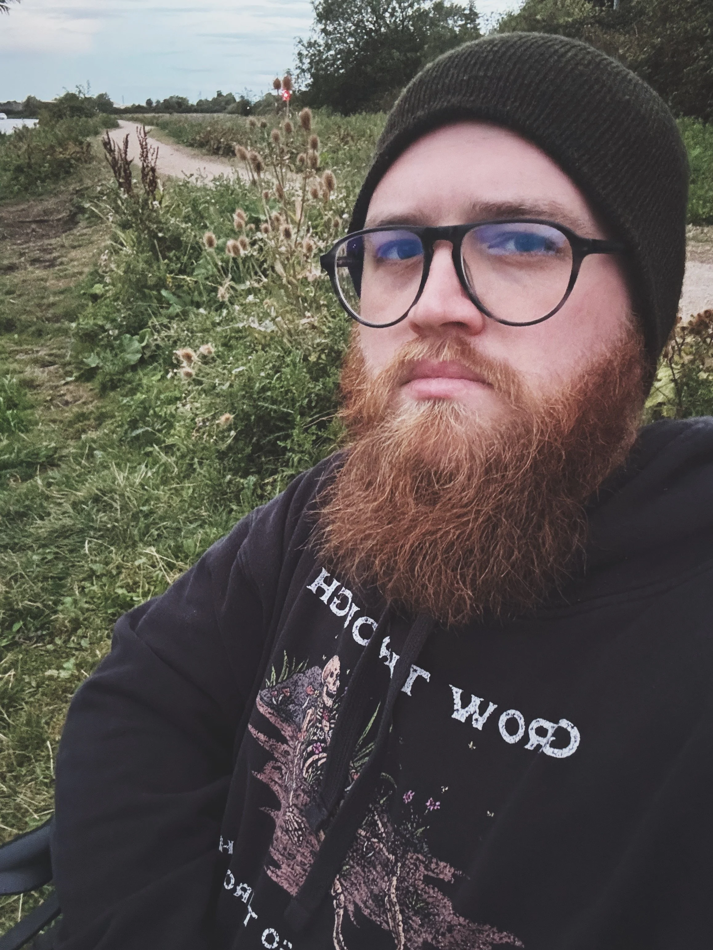 A man with a red beard and glasses, wearing a black beanie and hoodie, taking a selfie outdoors on a dirt path surrounded by wild plants and greenery, with a cloudy sky in the background.