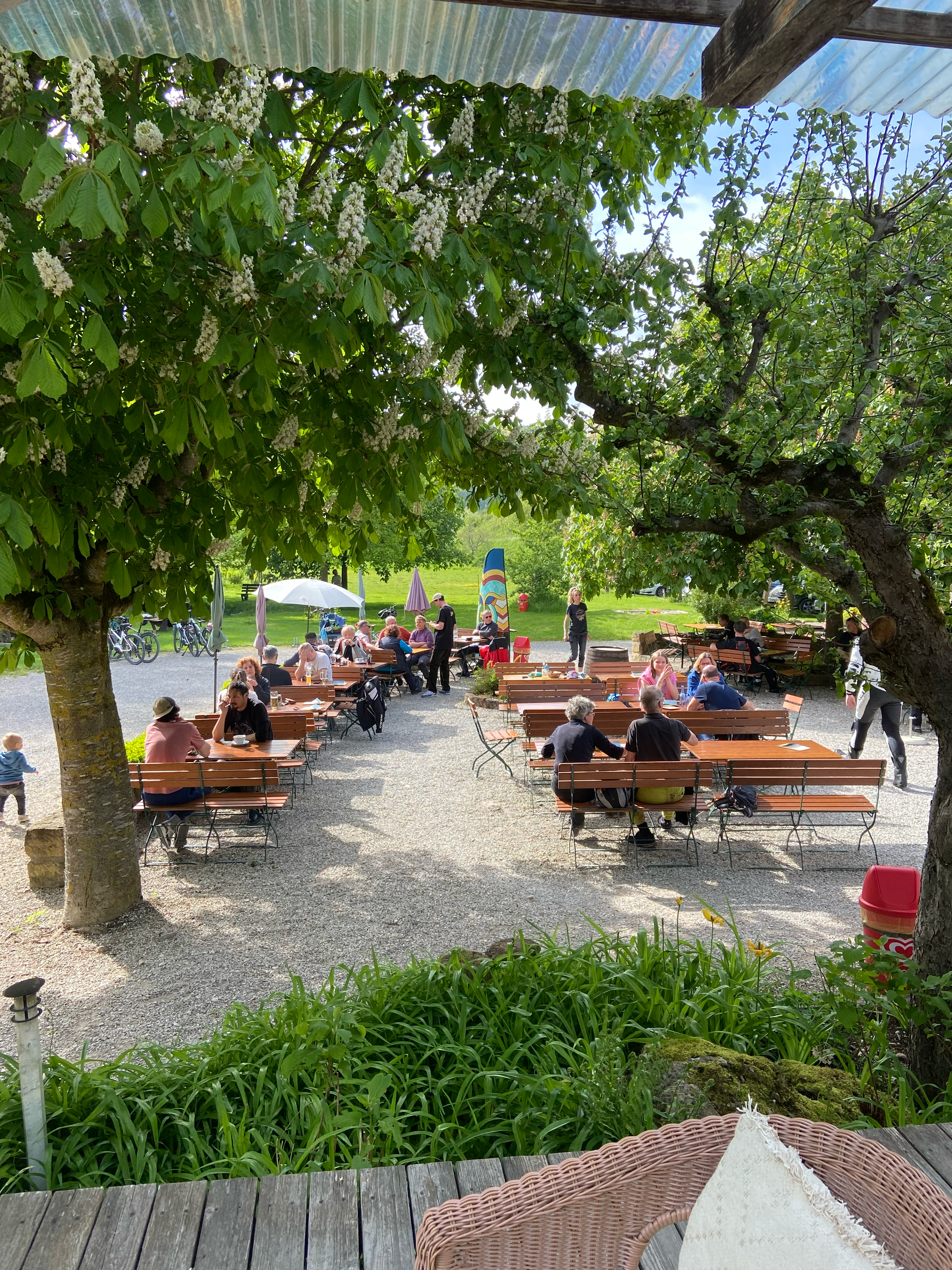 Menschen sitzen auf Bänken im Schatten unter Bäumen im Biergarten Bootshaus an der Lauter im Freien bei gutem Wetter, einige unter einem weißen Sonnenschirm.