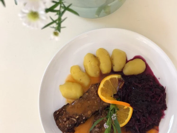 Ein Teller mit zwei Linsen-Nuss-Schnitten, Rotkohl, Kartoffeln, Orangenscheibe und einer kleinen grünen Pflanze, darüber ein Blumenstrauß mit weißen Blumen in einer Glasvase.