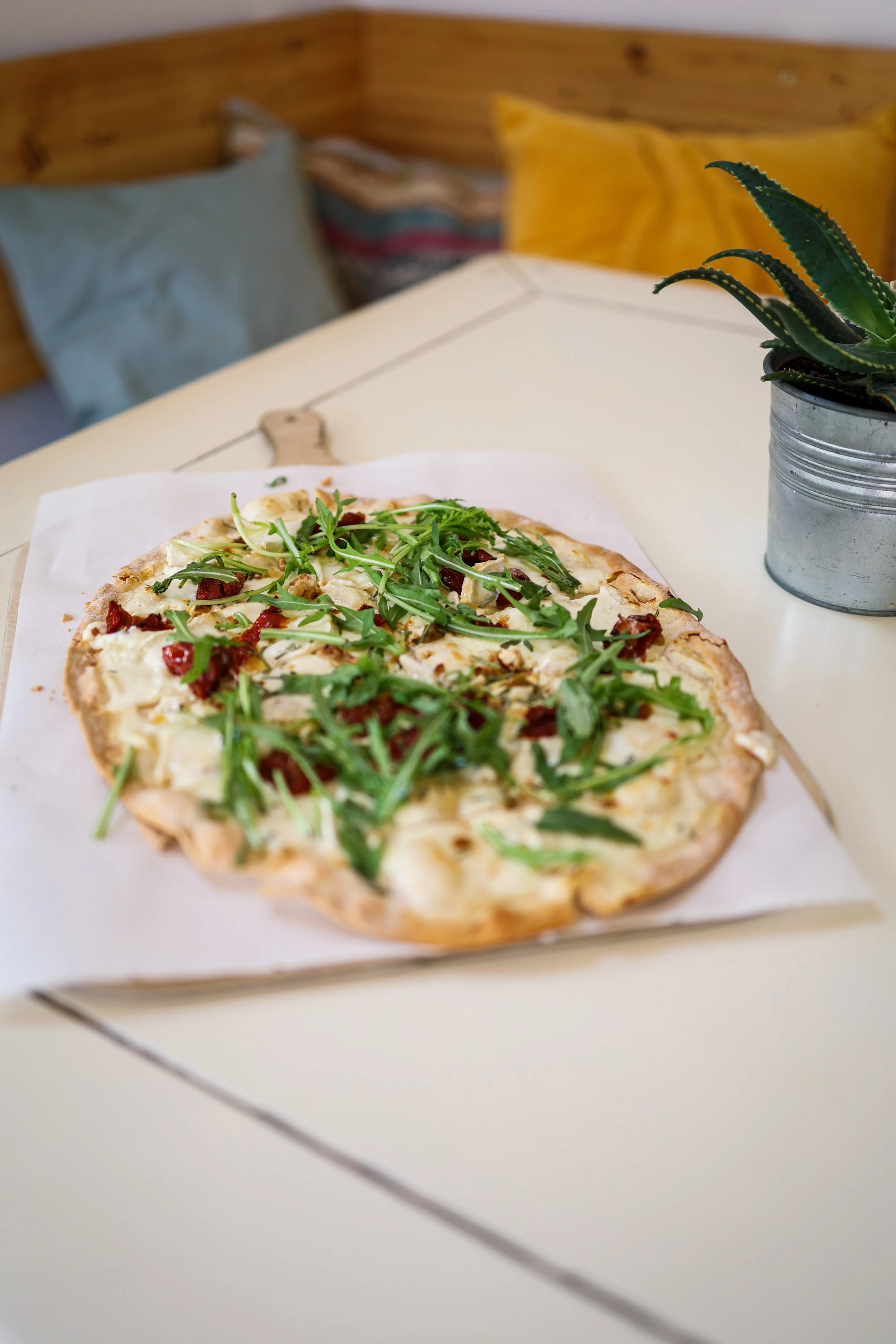 Ein zickiger Flammkuchen mit frischem Rucola, Tomatenstücken und Käse auf Creme Fraîche, steht auf einem Tisch neben einer Topfpflanze. Im Hintergrund sind bunte Kissen auf einer Eckbank. 