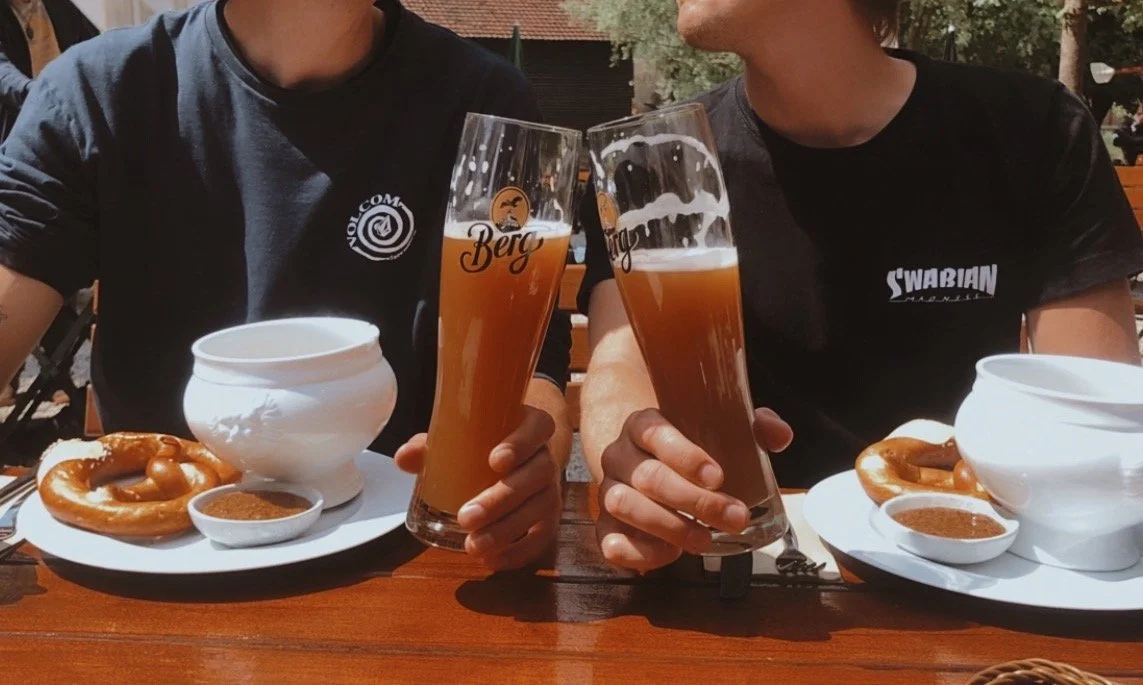 Zwei Menschen sitzen im Freien bei Weißwurstessen, trinken Bier und haben Brezeln auf dem Tisch.