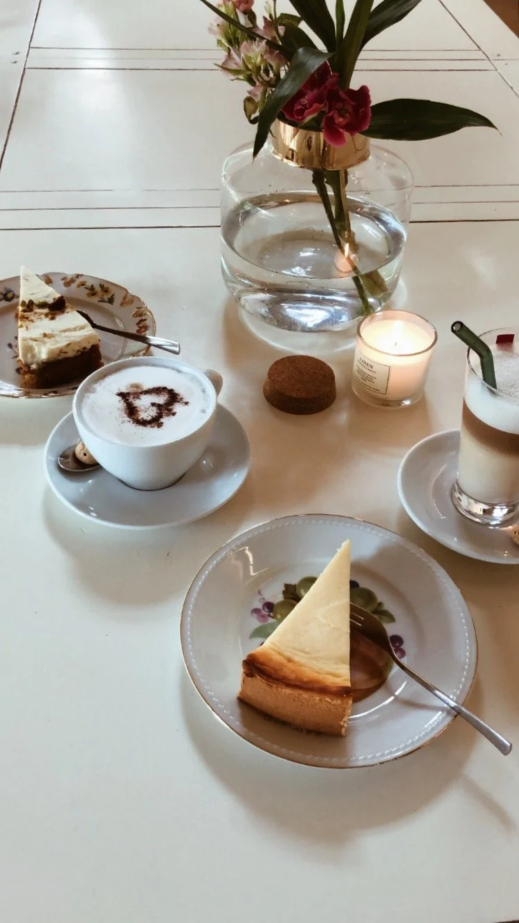 Tisch mit Blumen, einem Glas Wasser, einer Kerze, zwei Tassen Kaffee, einer Teetasse, einem Stück Käsekuchen und einem Stück Käsetorte.