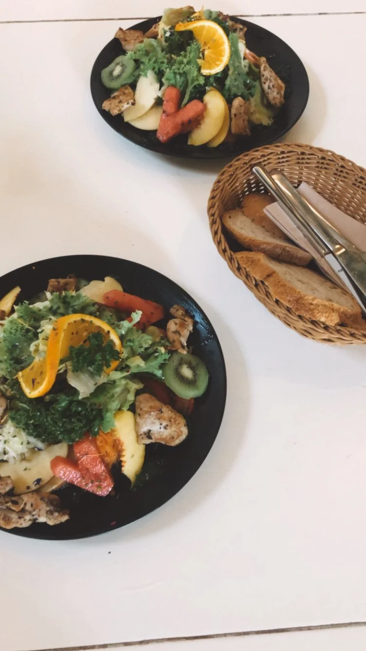 Zwei Teller mit bunter Salat mit Orangenscheiben, Kiwi, Tomaten, Gurken, gegrilltem Hähnchen, daneben ein Korb mit Bauernbrot auf einem weißen Tisch