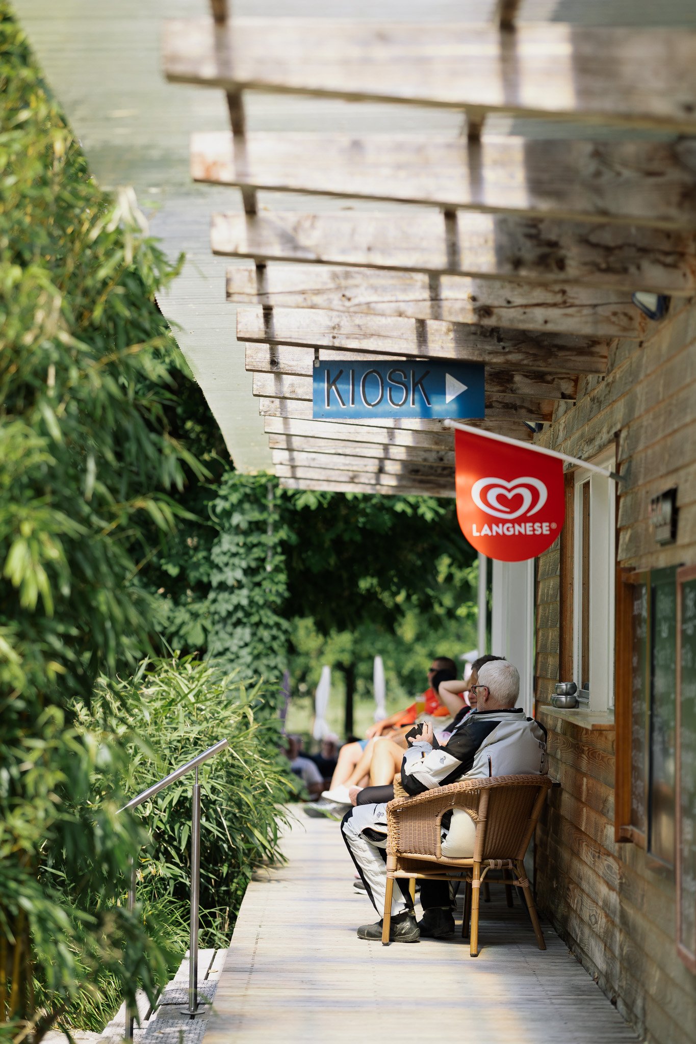 Menschen sitzen auf einer Veranda/Steg vor einem Holzgebäude, dem Bootshaus an der Lauter, mit einem roten Langnese-Werbebanner, ein blauer Kiosk-Schilder wird oben gezeigt, umgeben von grünen Büschen und Bäumen.