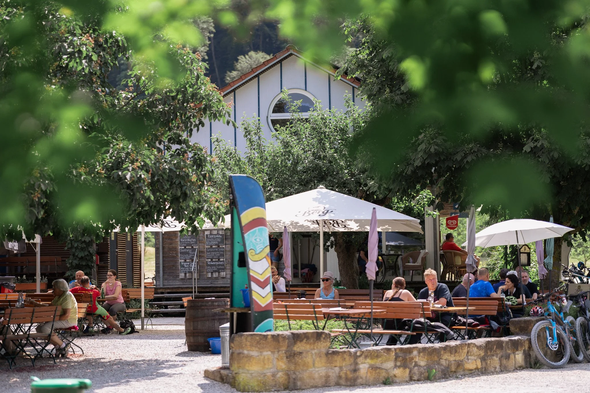 Aufnahme des Bootshaus an der Lauter mit mehreren Sitzbänken und Tischen, Menschen, die entspannt sitzen, Sonnenschirmen, Fahrrädern und Bäumen im Hintergrund.