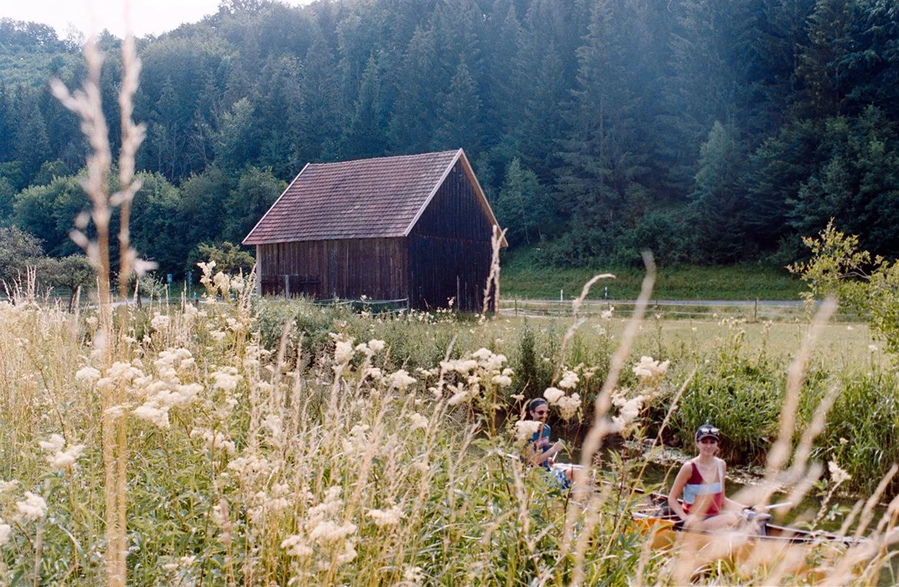 Zwei Mädchen in einem kleinen Boot auf einem Fluss, umgeben von hohen Gräsern und weißen Blumen, mit einem alten Holzhaus im Hintergrund, umgeben von Bäumen im Lautertal. 