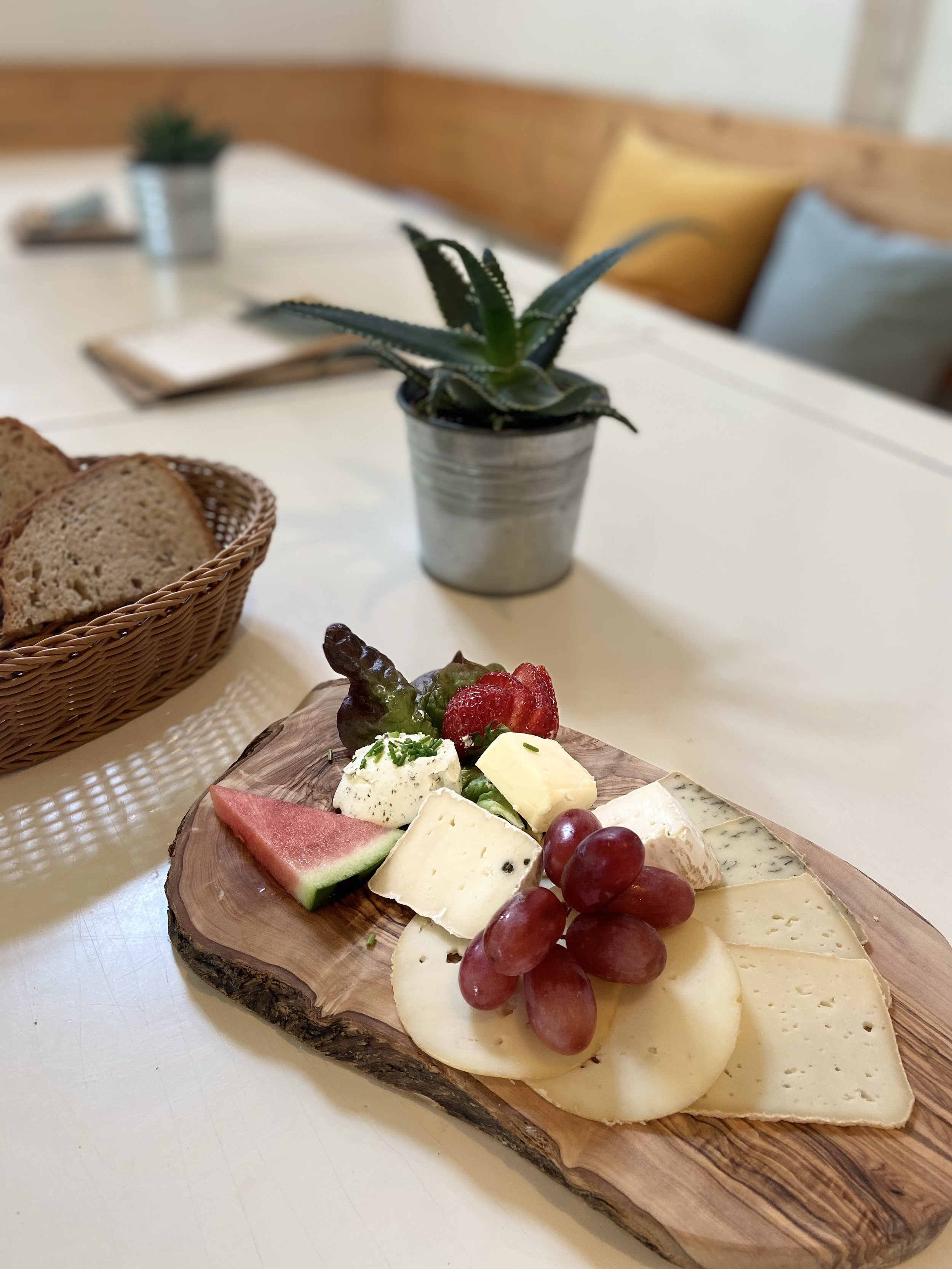 Käseplatte mit Trauben, Melone, Beeren und Kräutern auf Holzbrett, in der Nähe ein Korb mit Brot, im Hintergrund ein Topf mit Aloe Vera auf weißem Tisch.
