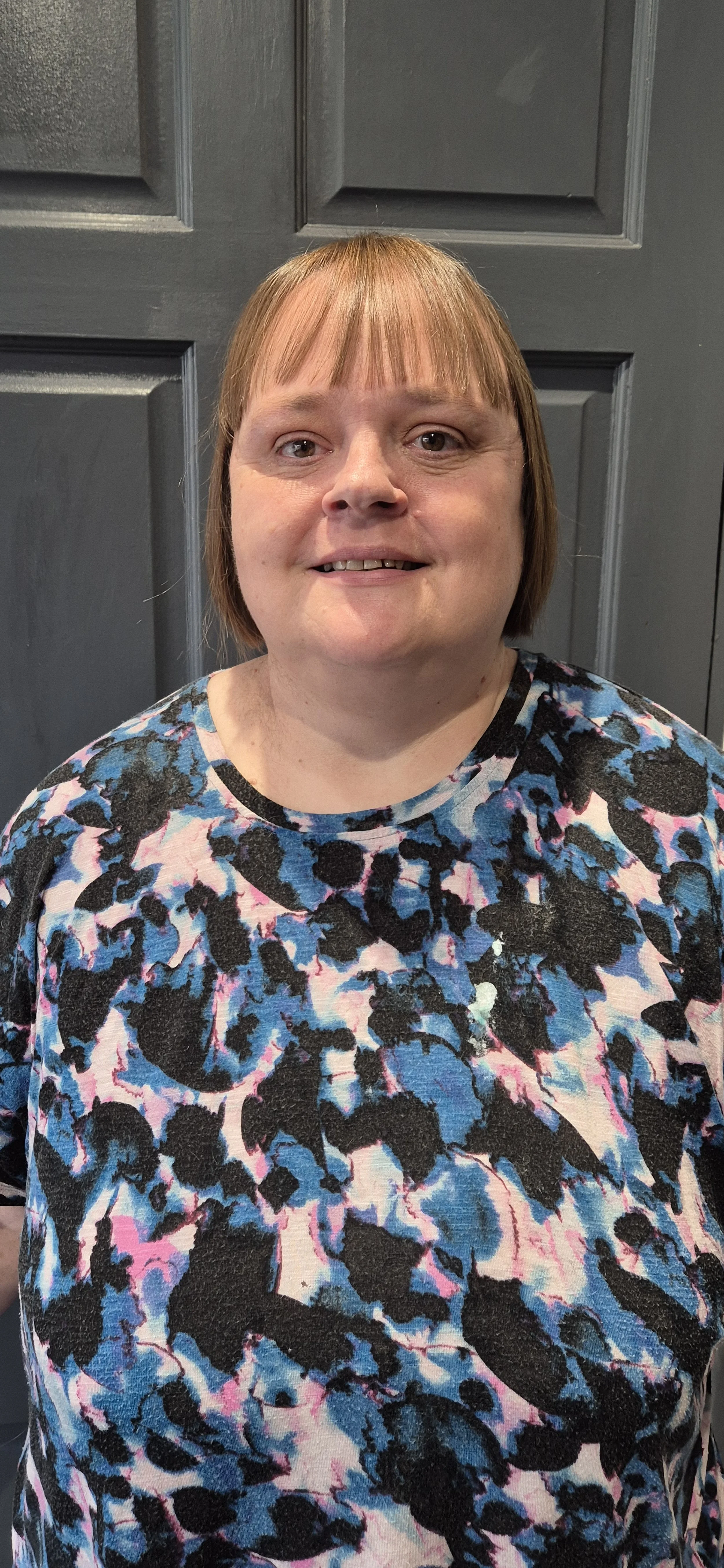 Portrait of Fiona Rudbach, founder of Rudbach Consultants. Standing in front of a black door wearing a blue and pink top. Fiona has shoulder length brown hair and brown eyes.