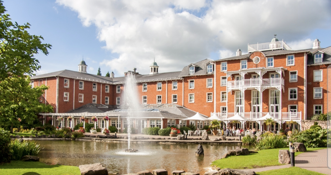 the back of the Alton Towers Hotel. This shows a lovely view of a lake with a working fountain and a 4 storey hotel behind. There is people sat outside having a drink on a sunny day.