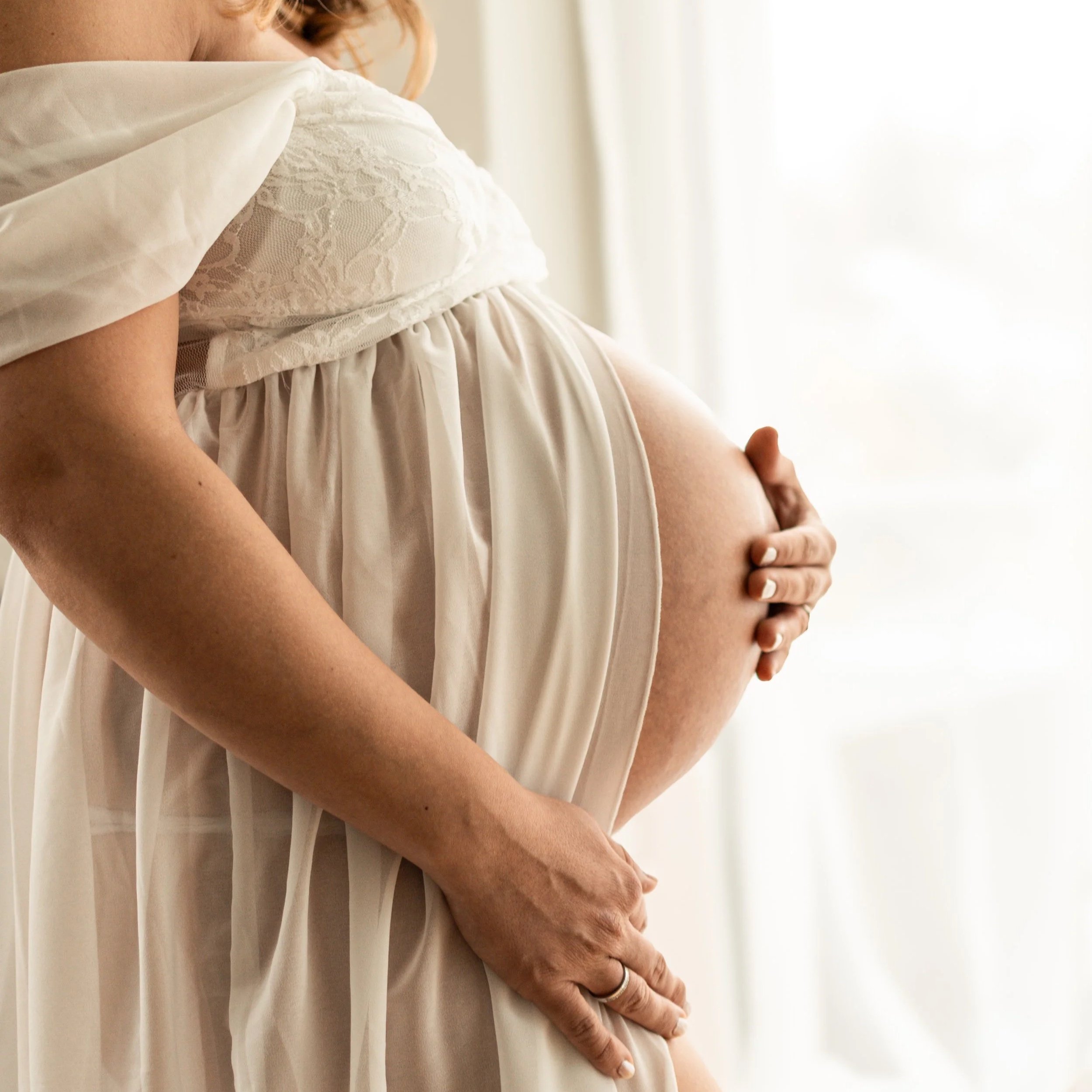 Eine schwangere Frau hält ihren Bauch mit beiden Händen, trägt ein fließendes, helles Kleid mit Spitze, vor einem hell erleuchteten Fenster.