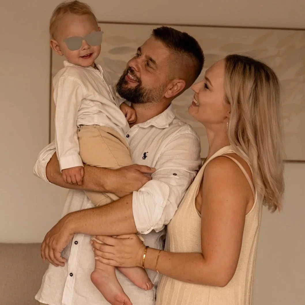 Familie mit Vater, Mutter und kleinem Kind, das lachend im Wohnzimmer spielt, Kind trägt Brille und Sonnenbrille.