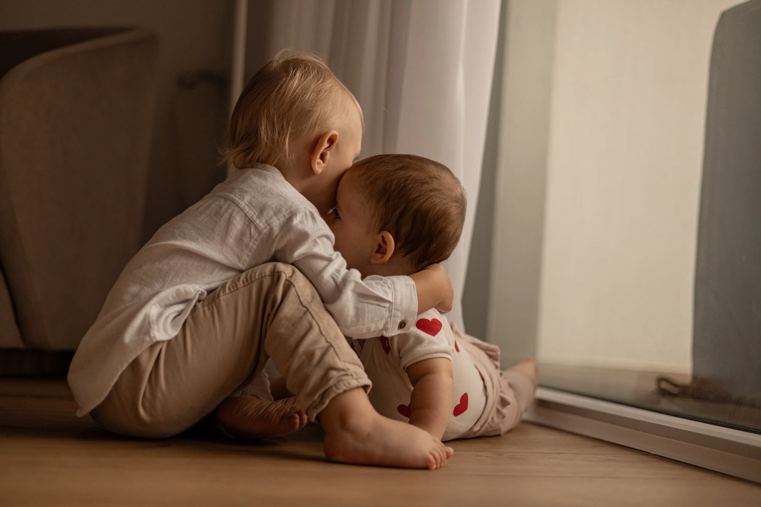 Zwei kleine Kinder, eines sitzt auf dem Boden und der andere liegt auf seinem Schoß, küssen sich liebevoll vor einer Glas-Tür, im Raum mit weichem Licht.