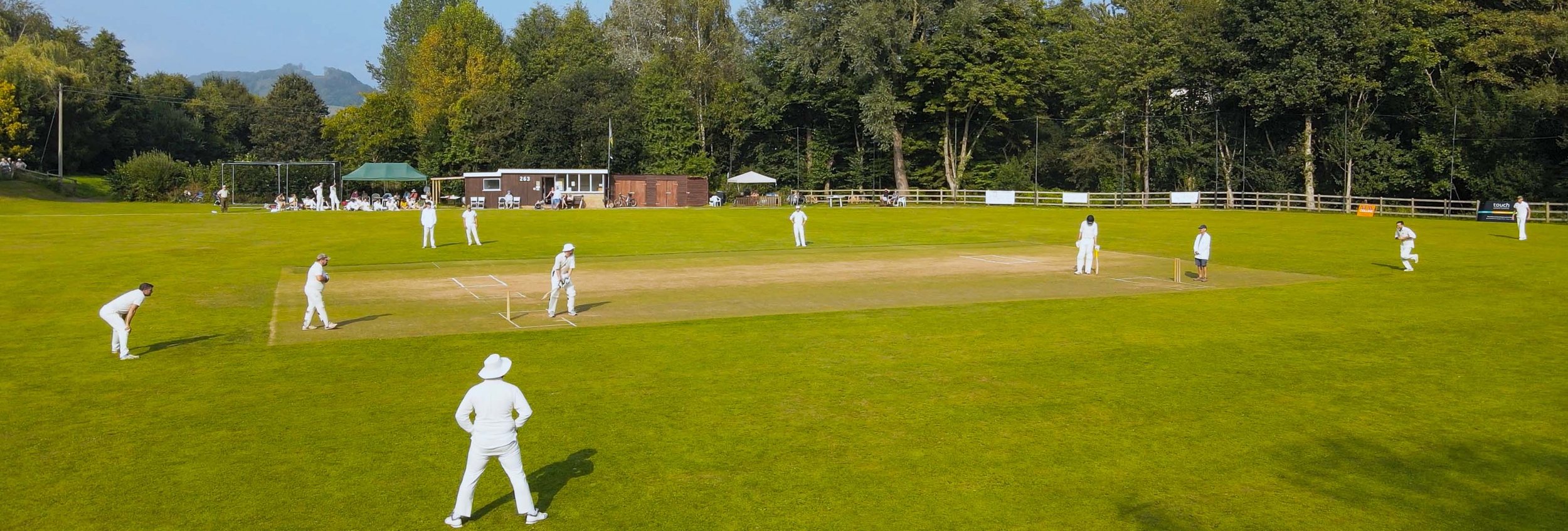 A cricket match is being played on a grassy field with trees in the background. Several players dressed in white are on the field, some in ready positions. There are spectators and facilities in the background.