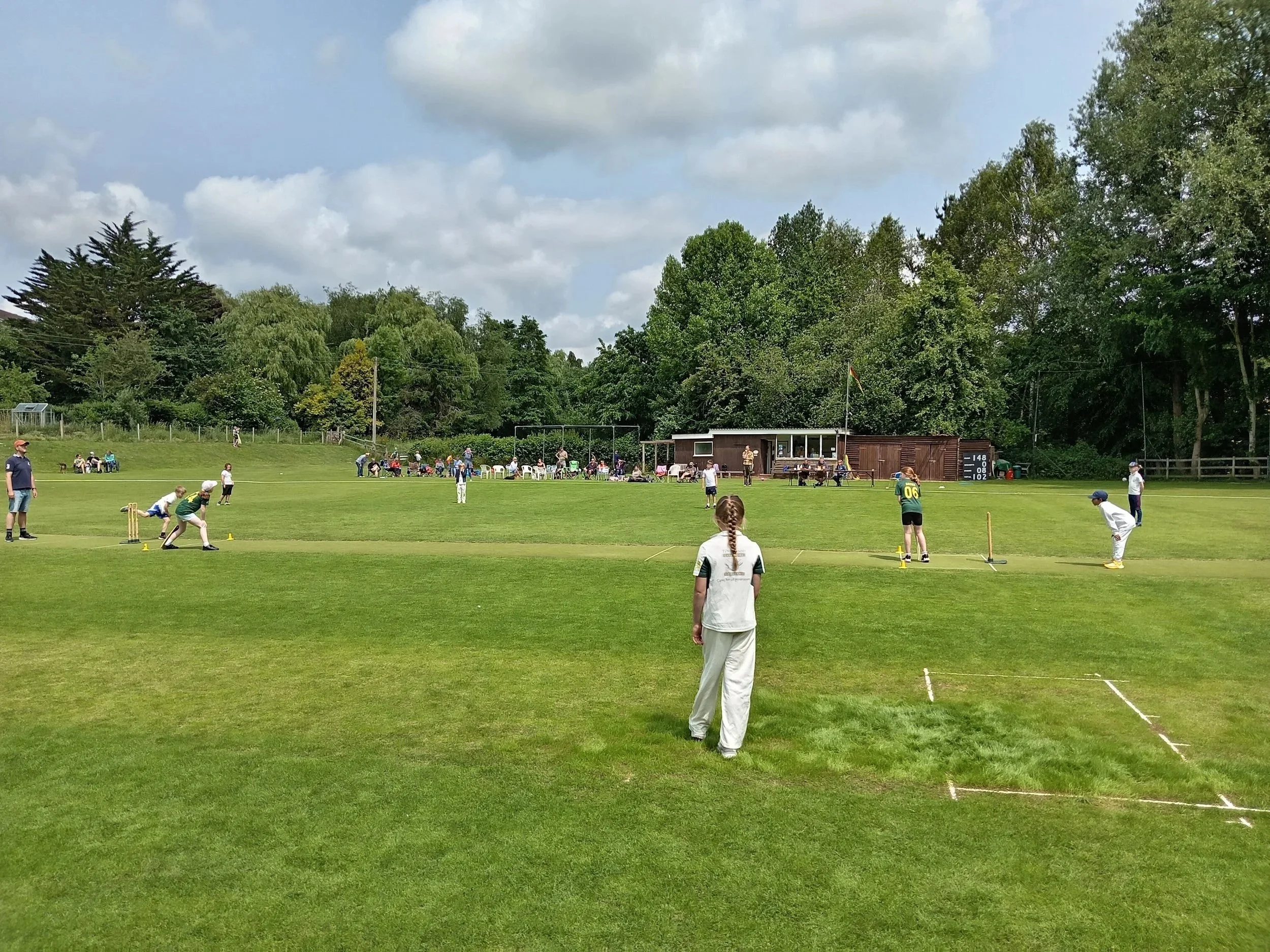 Children playing cricket on a lush green field with spectators in the background on a cloudy day.