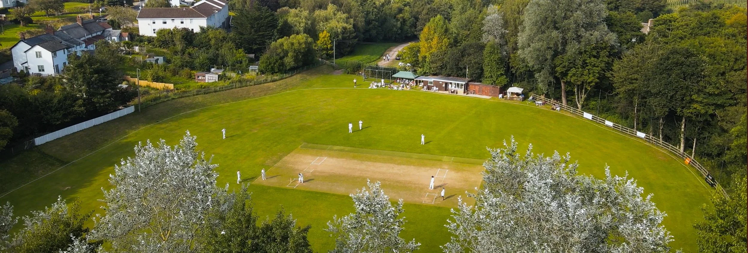 An aerial view of a cricket field with players on the pitch and along the boundary, adjacent to a fenced grassy area and a forested area with trees, and a small building with a covered seating area on the side.