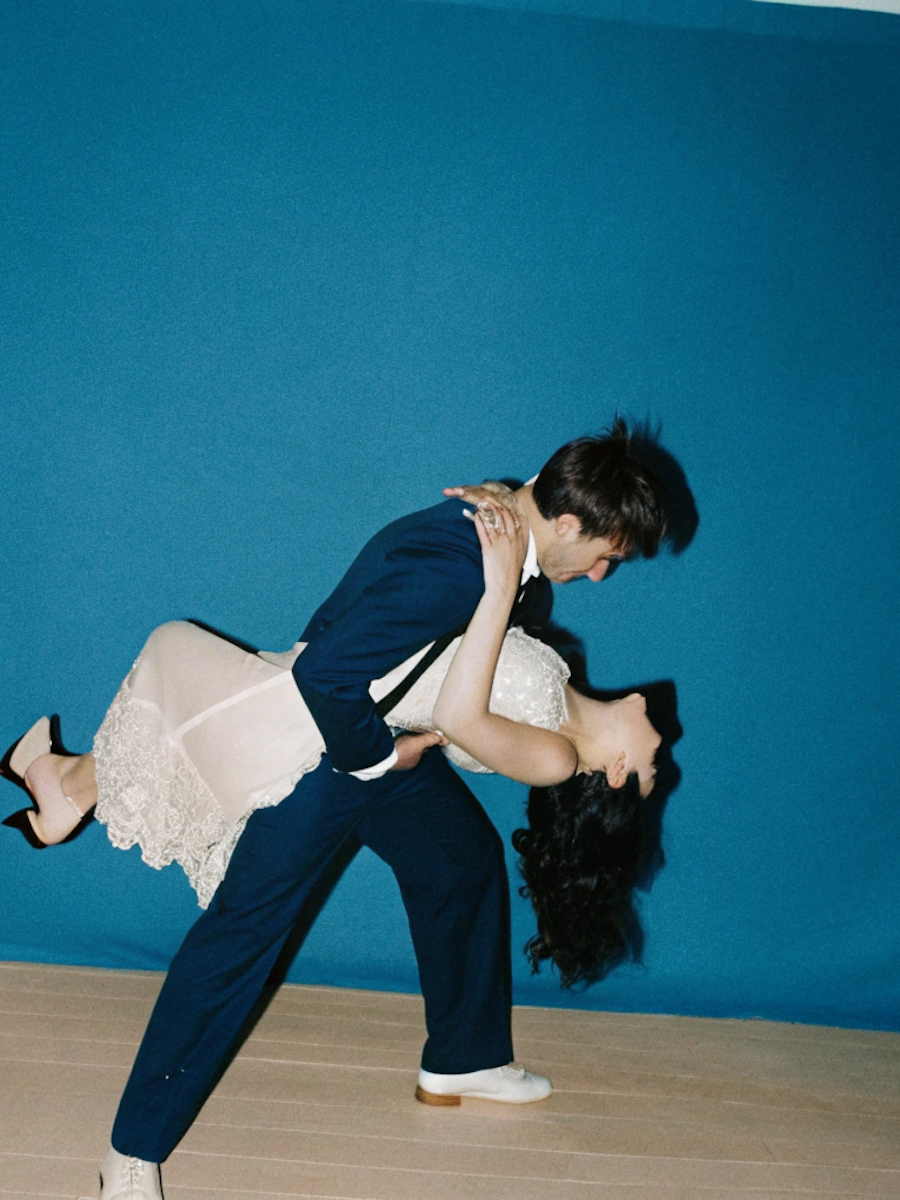 A man in a dark suit and a woman in a white dress are dancing, with the woman being dipped backwards in front of a blue wall.