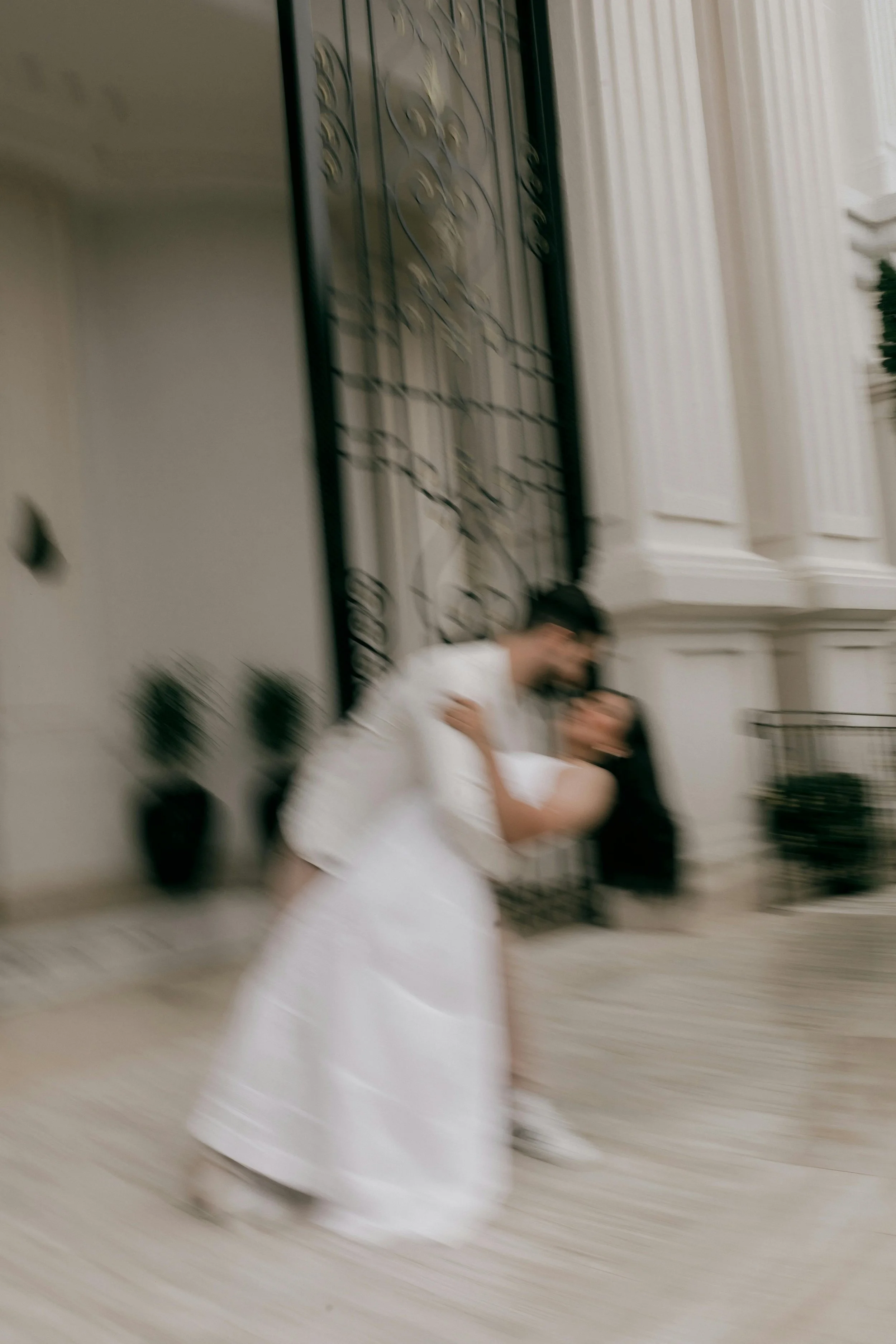 A blurred photo of a couple dancing in an elegant setting with white architectural elements and potted plants.