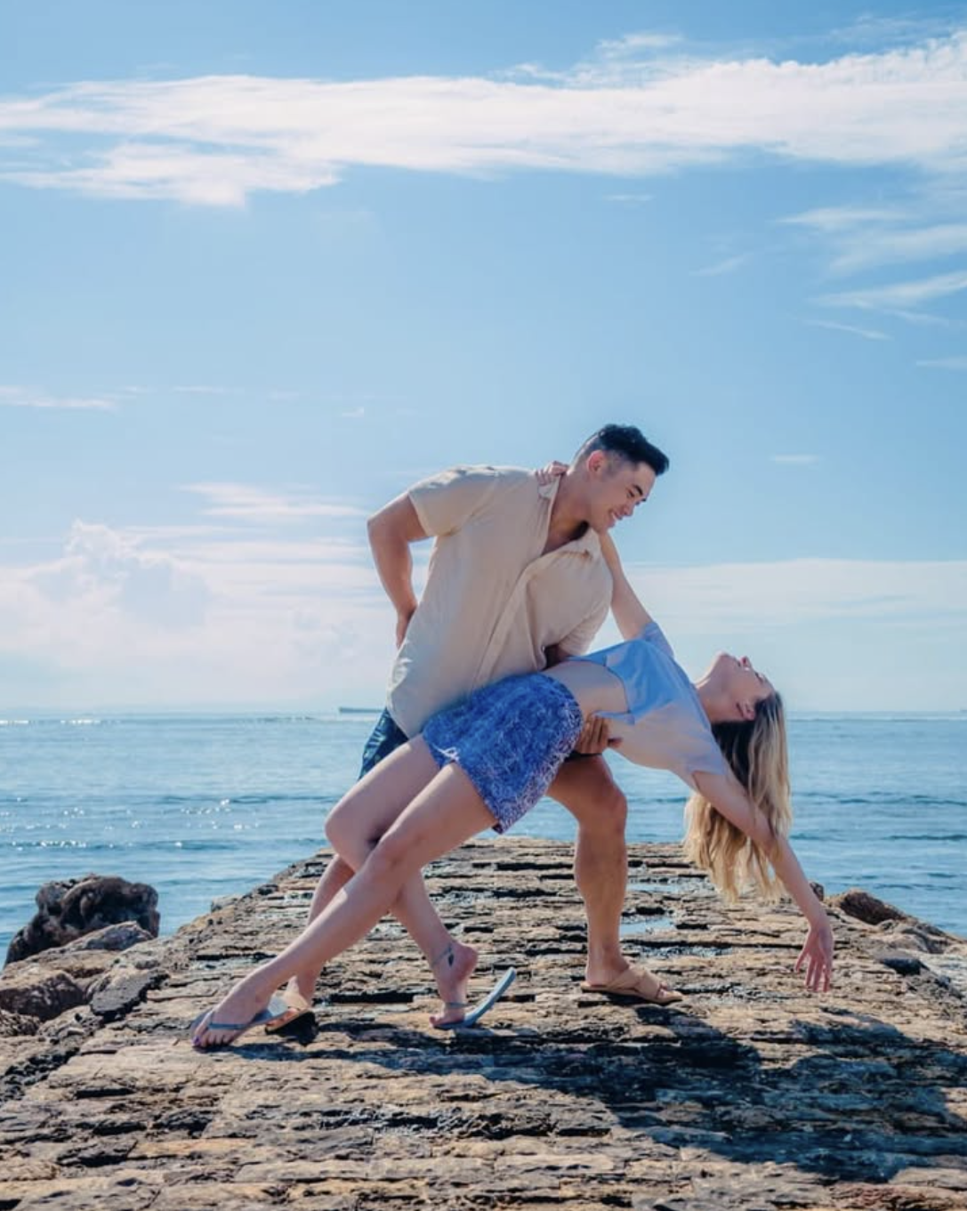 A couple engaged in a playful wrestling match on a rocky dock at the beach during daytime, with the ocean and a blue sky with clouds in the background.