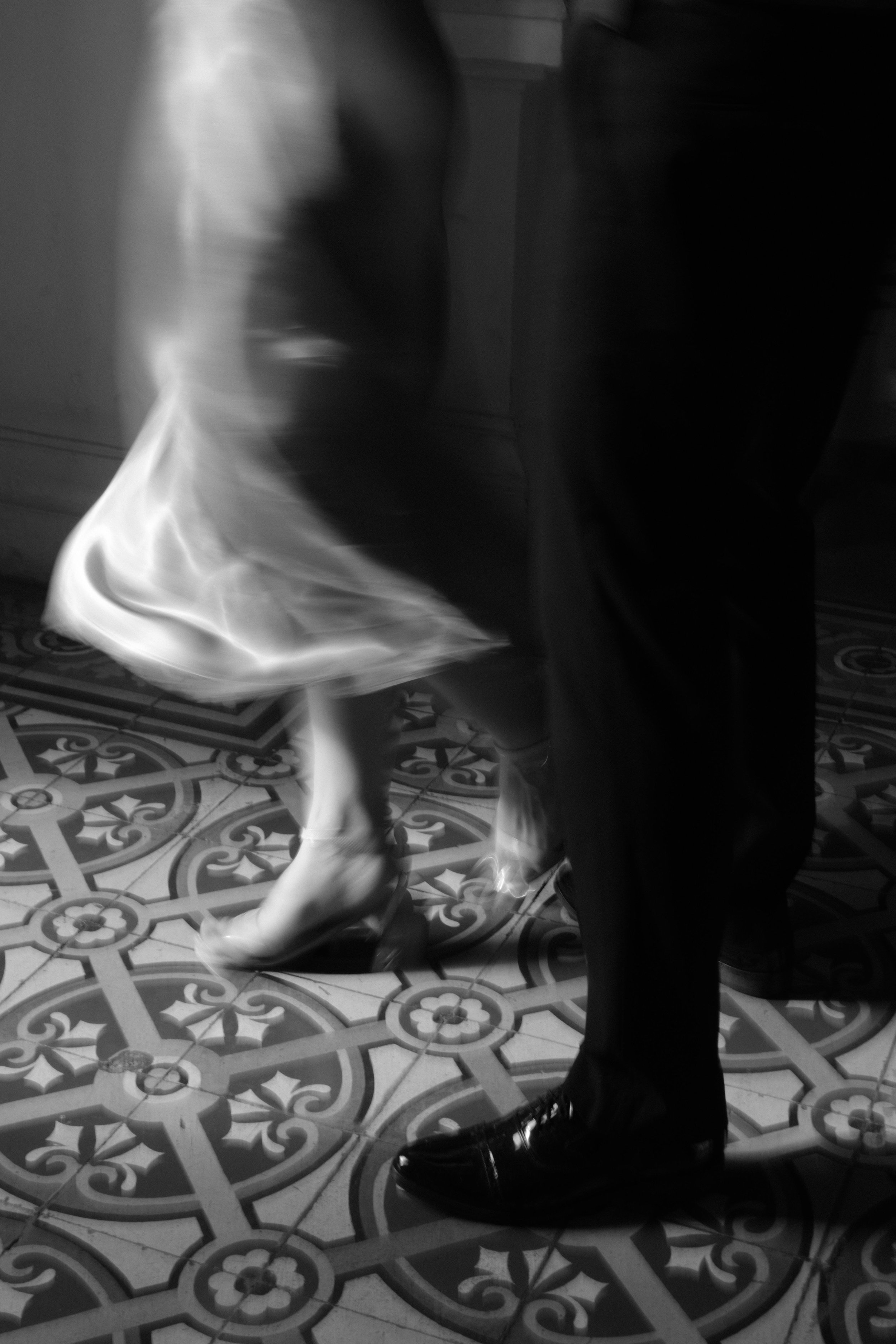 Black and white photo of a dancing couple, focusing on their legs and shoes, with intricate patterned floor tiles visible.