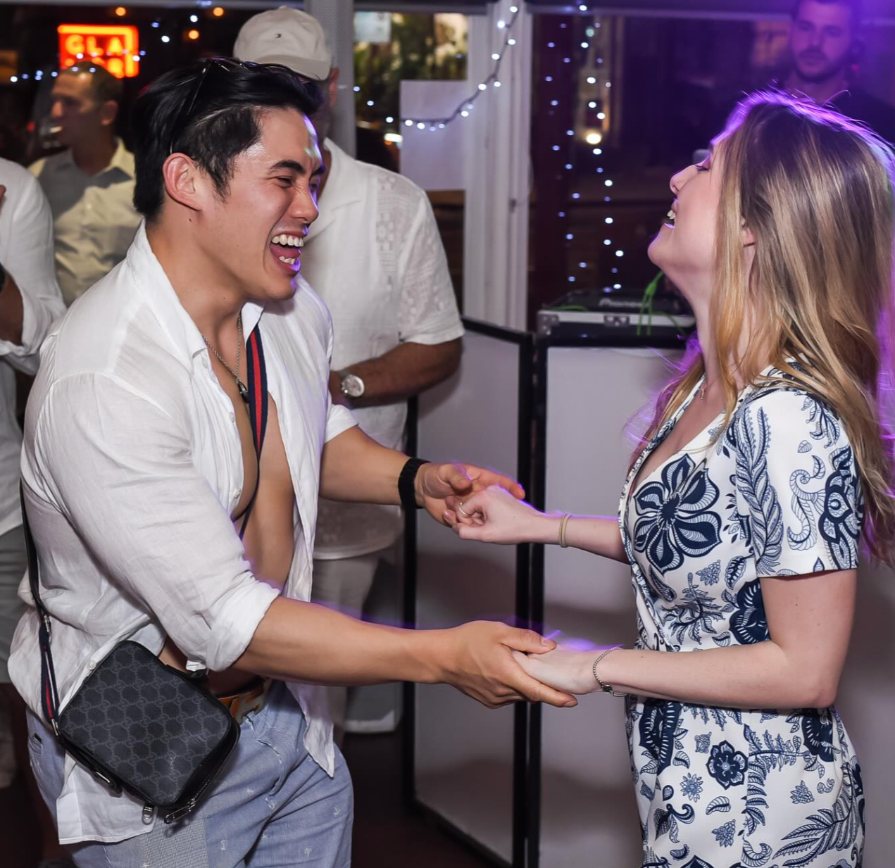 Two people, a man and a woman, are holding hands and laughing at each other at a social gathering. There are other people in the background and festive string lights.