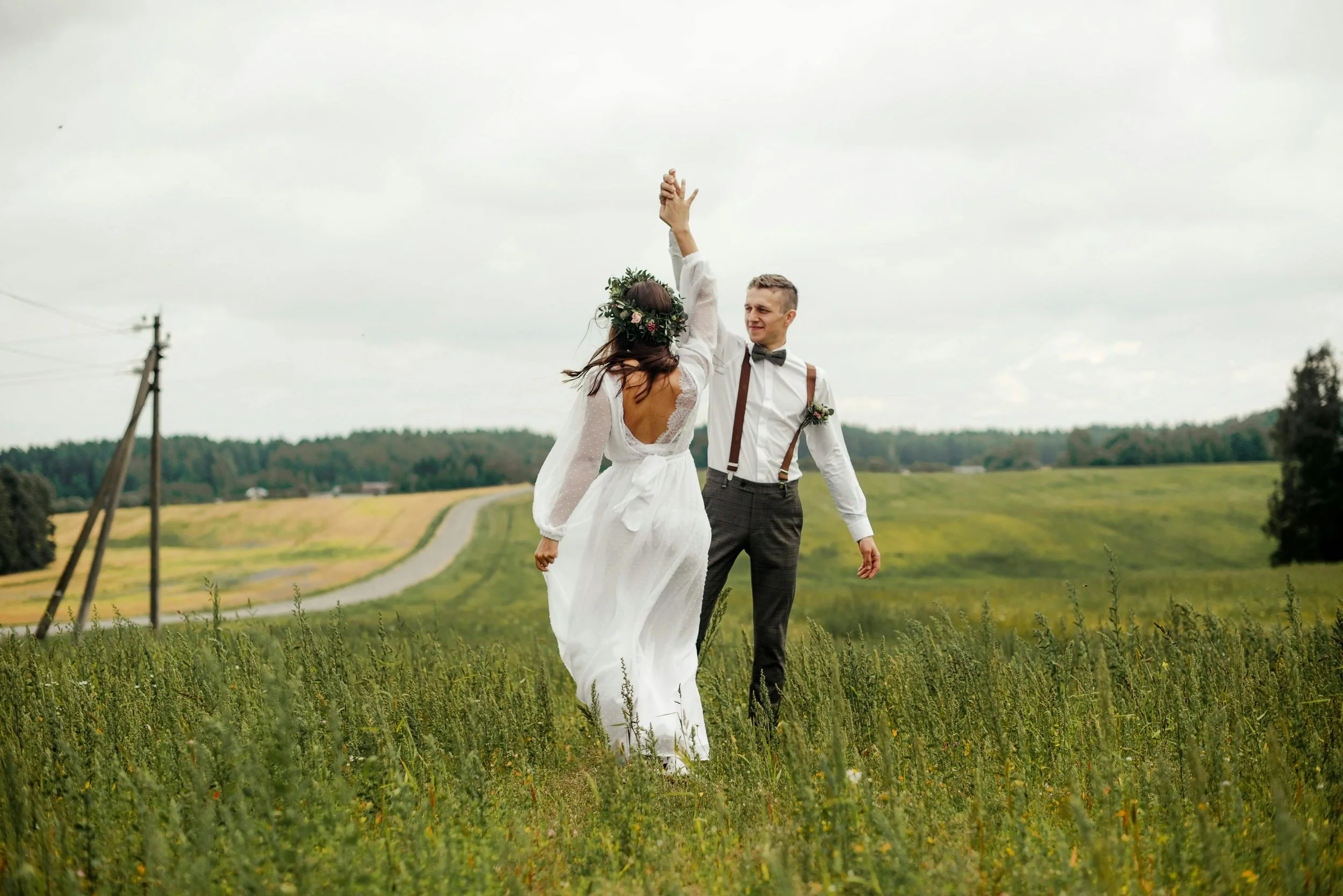 A couple dancing in a field on their wedding day, the bride wearing a white dress and floral crown, the groom in a tuxedo with suspenders, holding hands with the bride.