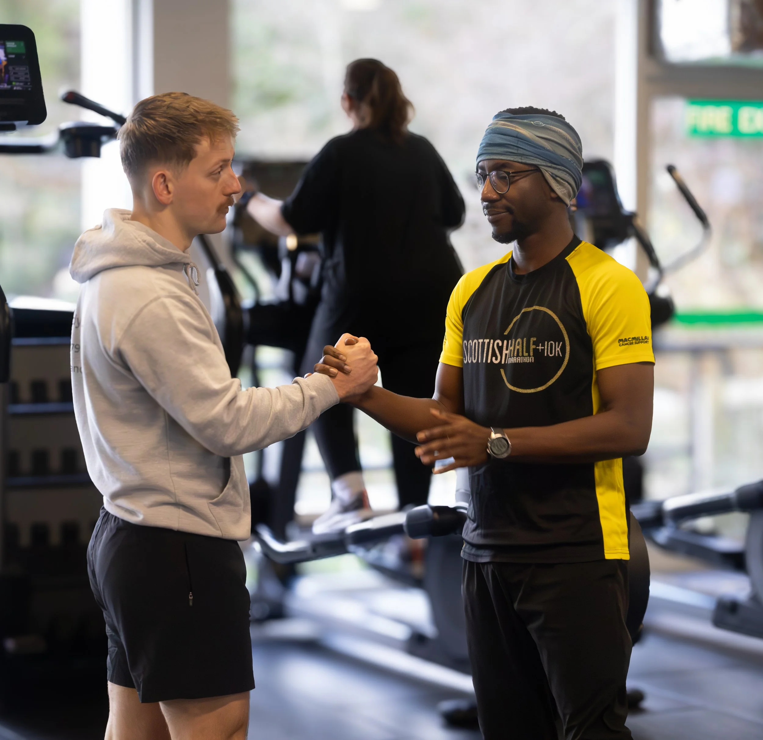 Two men shaking hands in a gym, with exercise equipment and a woman on a treadmill in the background.