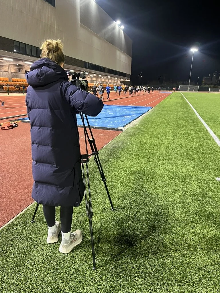 A person wearing a dark coat and white sneakers is filming or photographing a group of runners on a track at night, with bright stadium lights illuminating the scene.