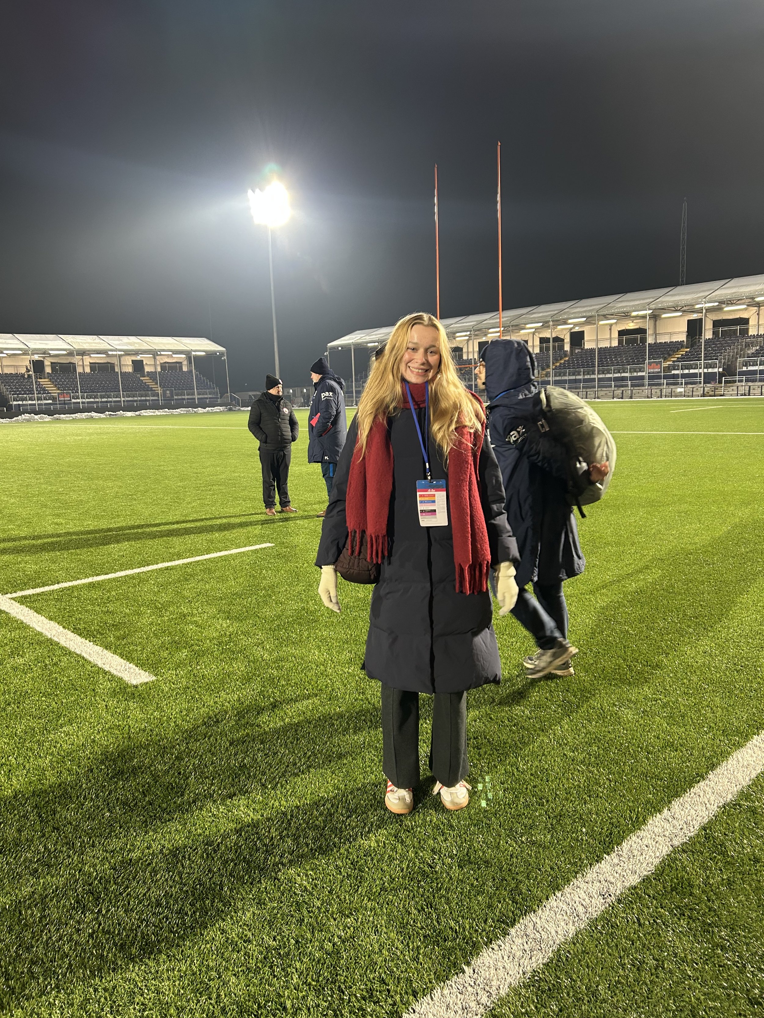 A woman standing on a football field at night, wearing a long coat and red scarf, smiling at the camera, with stadium seating and snow in the background.