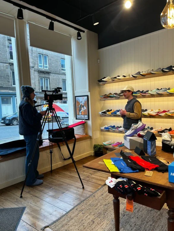 Person filming in a shoe store with shelves of sneakers on the wall, during a TV interview or recording.