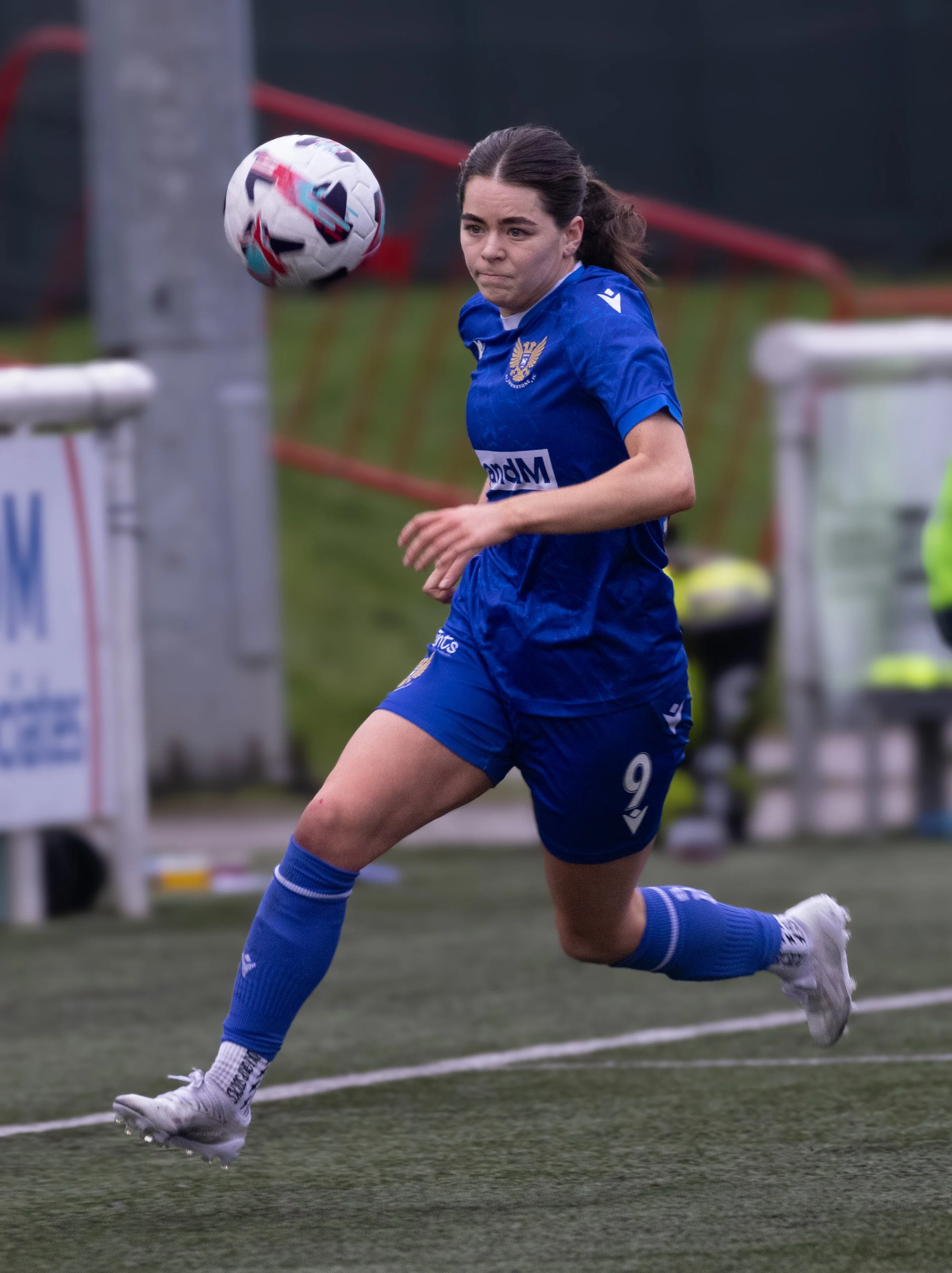 A female soccer player in a blue uniform running on the field with a soccer ball nearby, during a game.