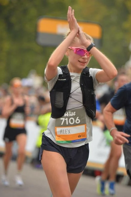A young woman in athletic gear, wearing sunglasses and a bib number 7106 with the name Alex, participating in a marathon or running event, crossing the finish line with her hands together in a celebratory gesture.