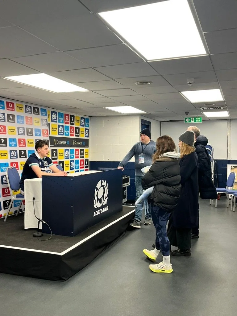 A young male athlete seated at a press conference table with a Scotland sports logo, facing a small group of reporters and fans in a room decorated with rugby and sports sponsorship banners.